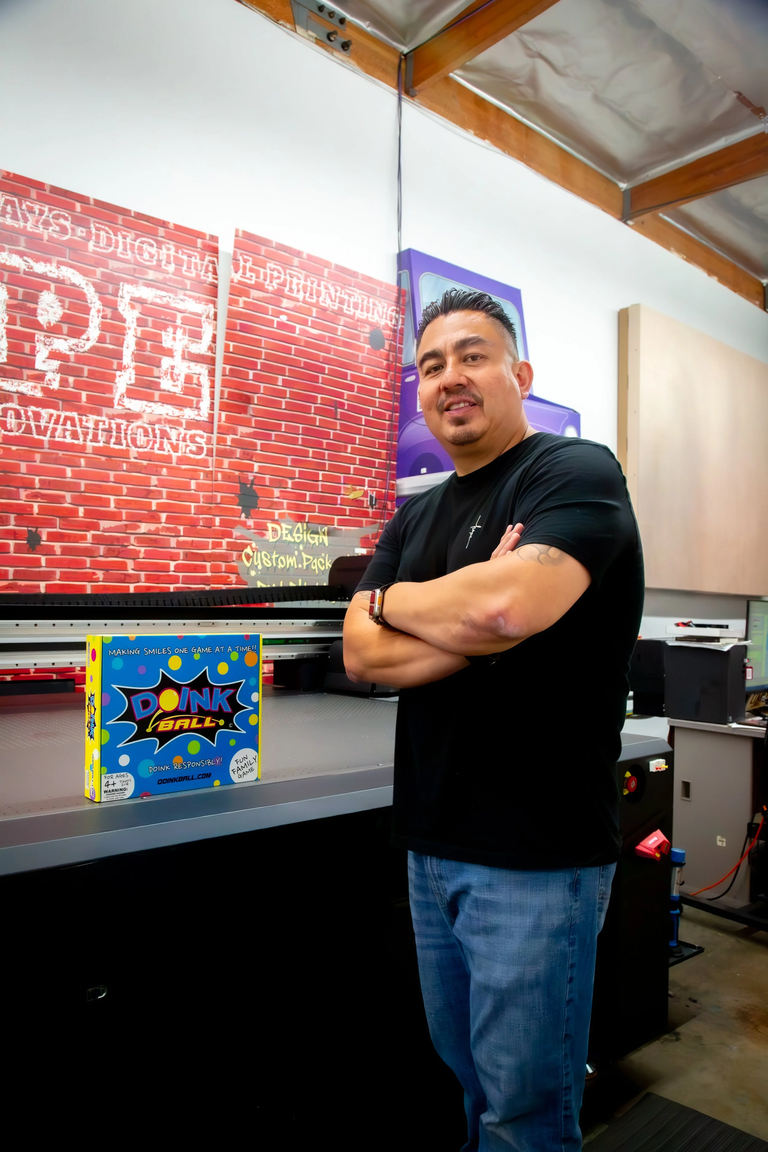 A man stands with arms crossed in front of a ping pong ball game display that reads "PING BALL" and a brick wall mural background with neon sign text.