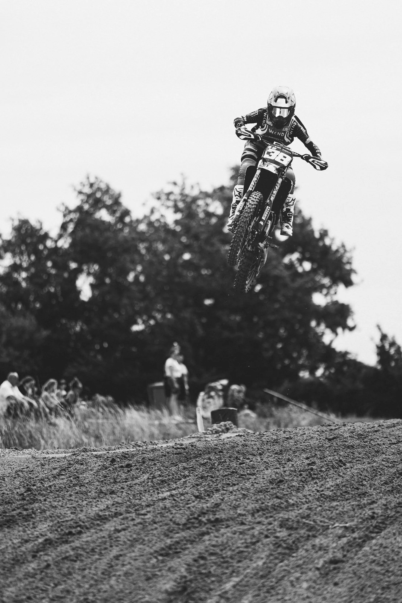 Een motocrossrace met een motorrijder die in de lucht springt boven een zandbaan, met een groep toeschouwers op de achtergrond.