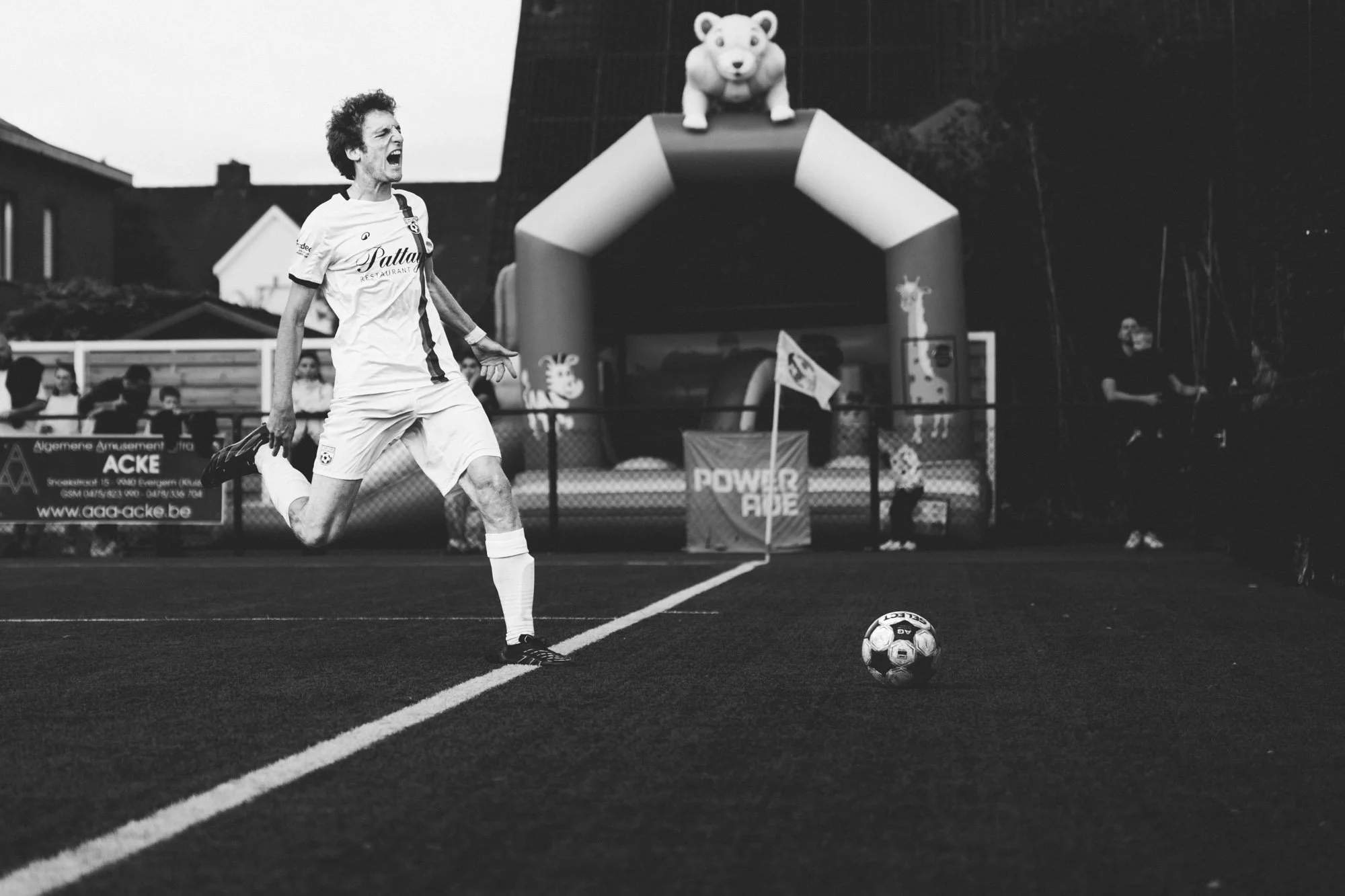 Een man in een voetbaloutfit die schreeuwt en zich voorbereidt om een penalty te nemen, met een voetbal voor zich, op een voetbalveld tijdens een jeugdwedstrijd, met mensen en een grote kleurrijke speeltoestel op de achtergrond.
