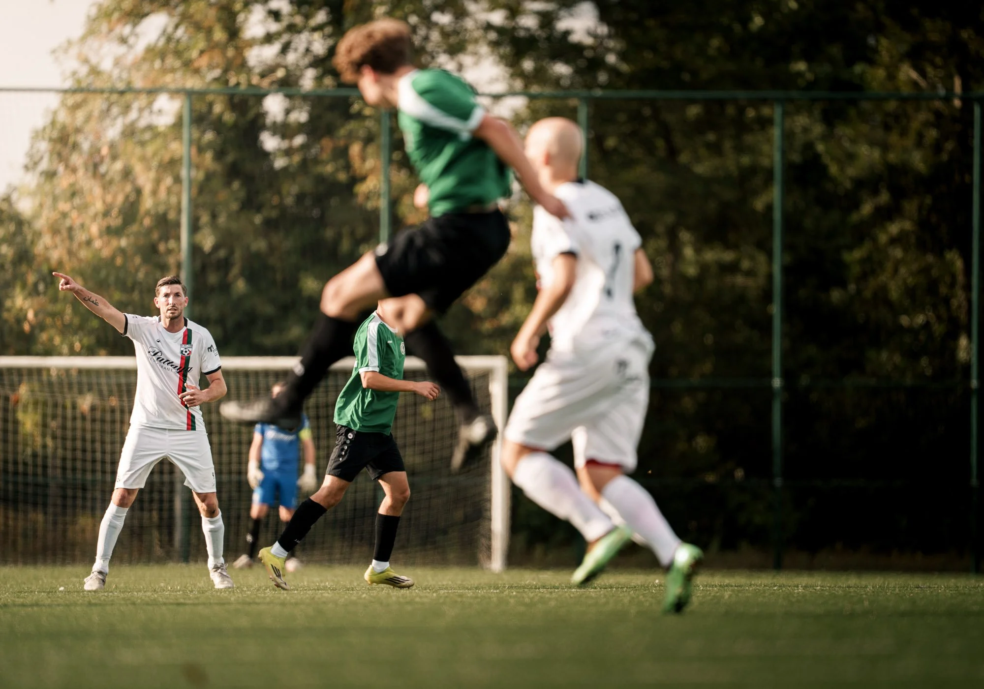 Mannen in een wit voetbal tenue met zwarte en rode strepen kijkt naar de bal, terwijl jongens in groene en witte truien betrokken zijn bij een voetbalwedstrijd op een grasveld.