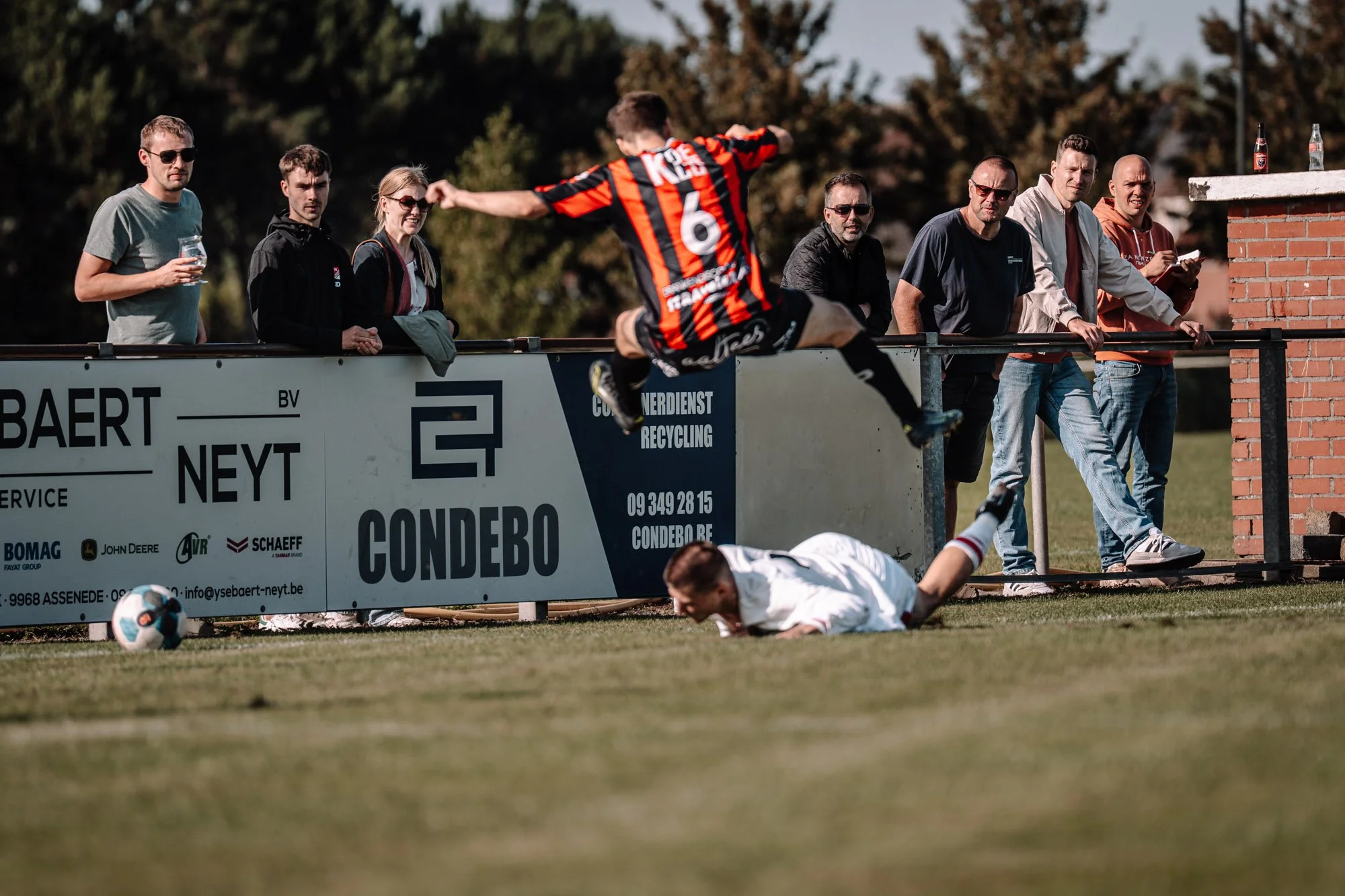 Speler die op de grond ligt na een botsing tijdens een voetbalwedstrijd, met toeschouwers die toekijken vanaf de zijlijn.