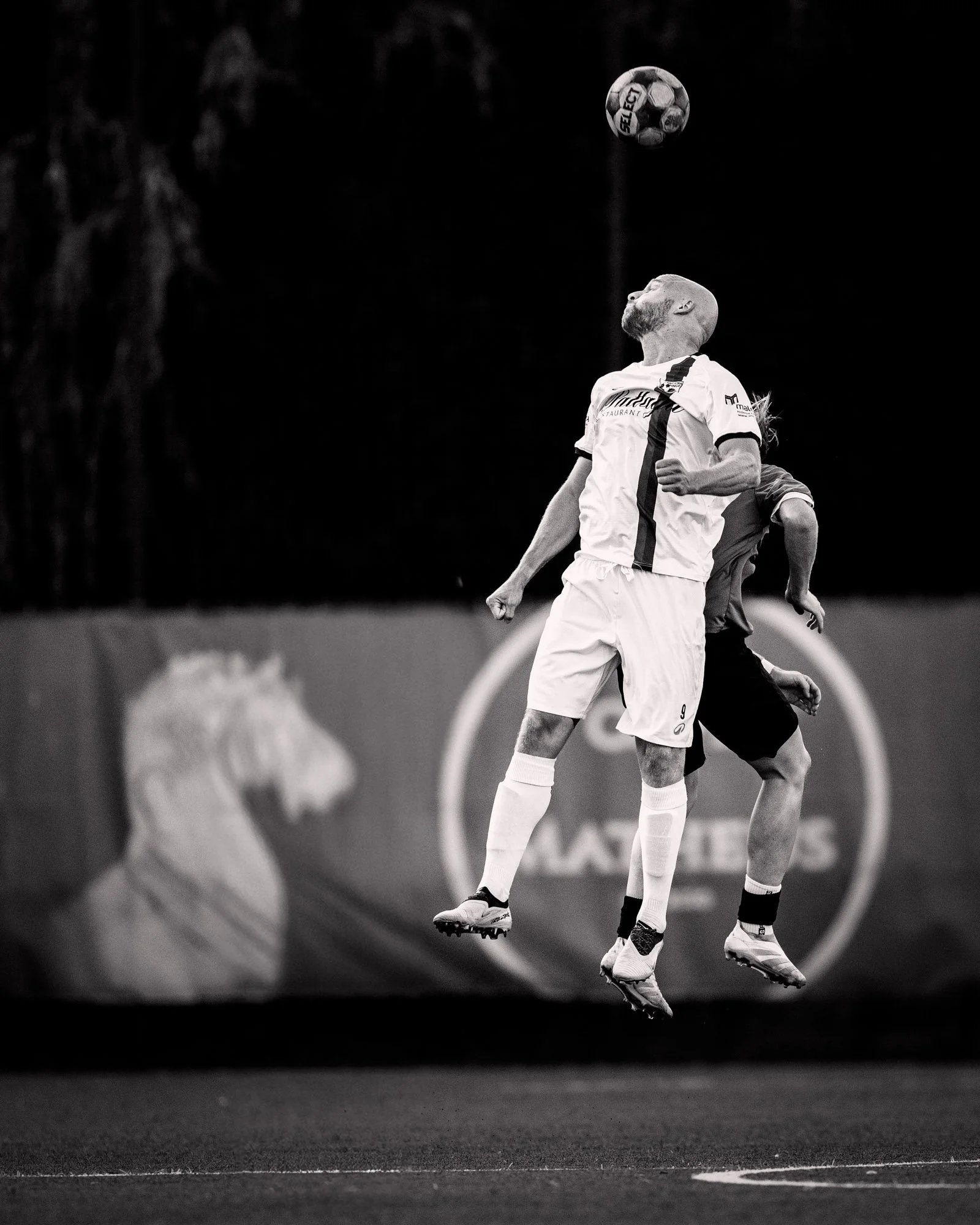 Zwart-wit foto van twee mannen die tegelijk springen en een bal omhoog werpen op een sportveld.