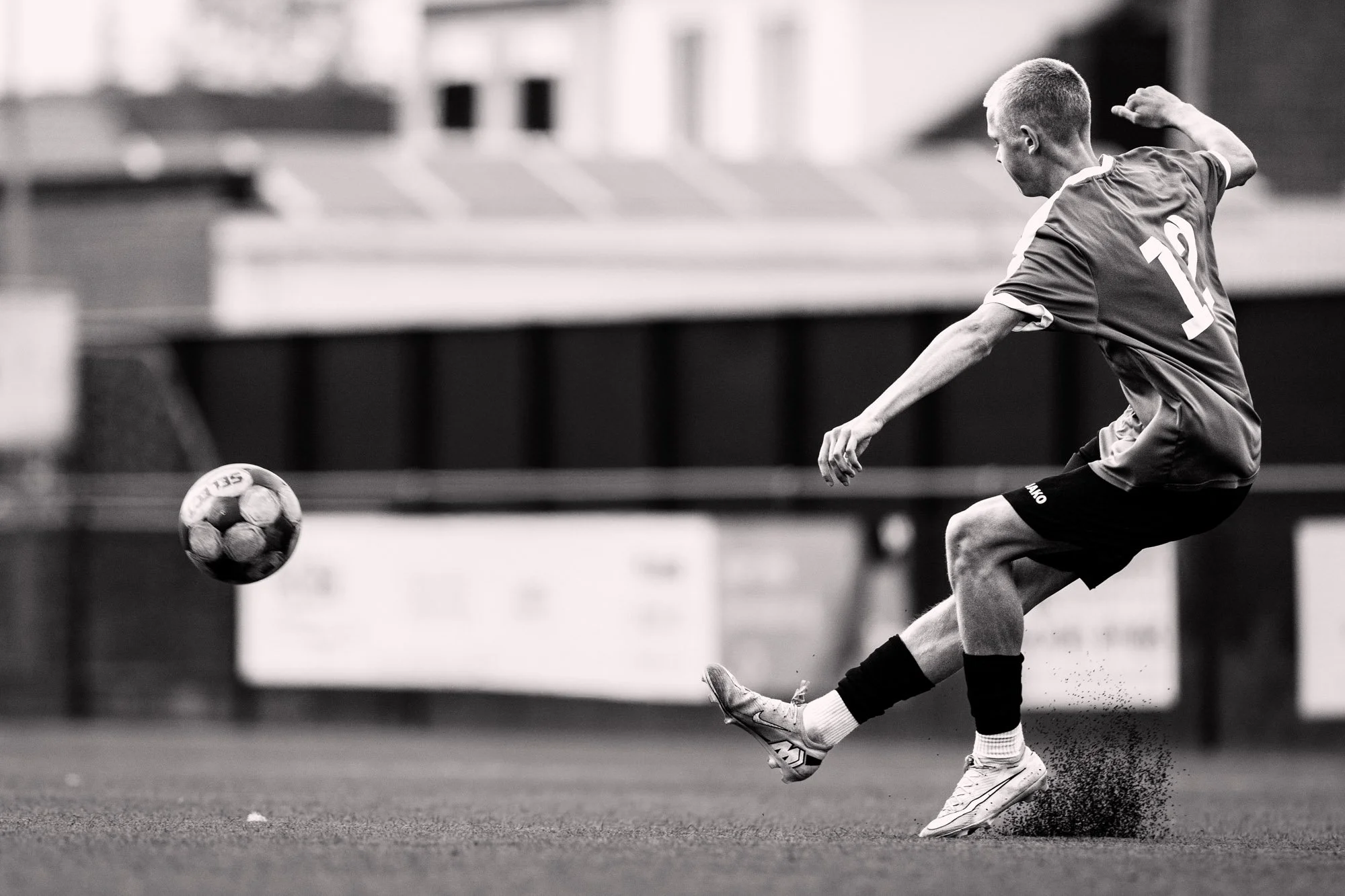 Zwart-witfoto van een man die net een voetbal trapt op een grasveld, met een sportoutfit en korte haren.
