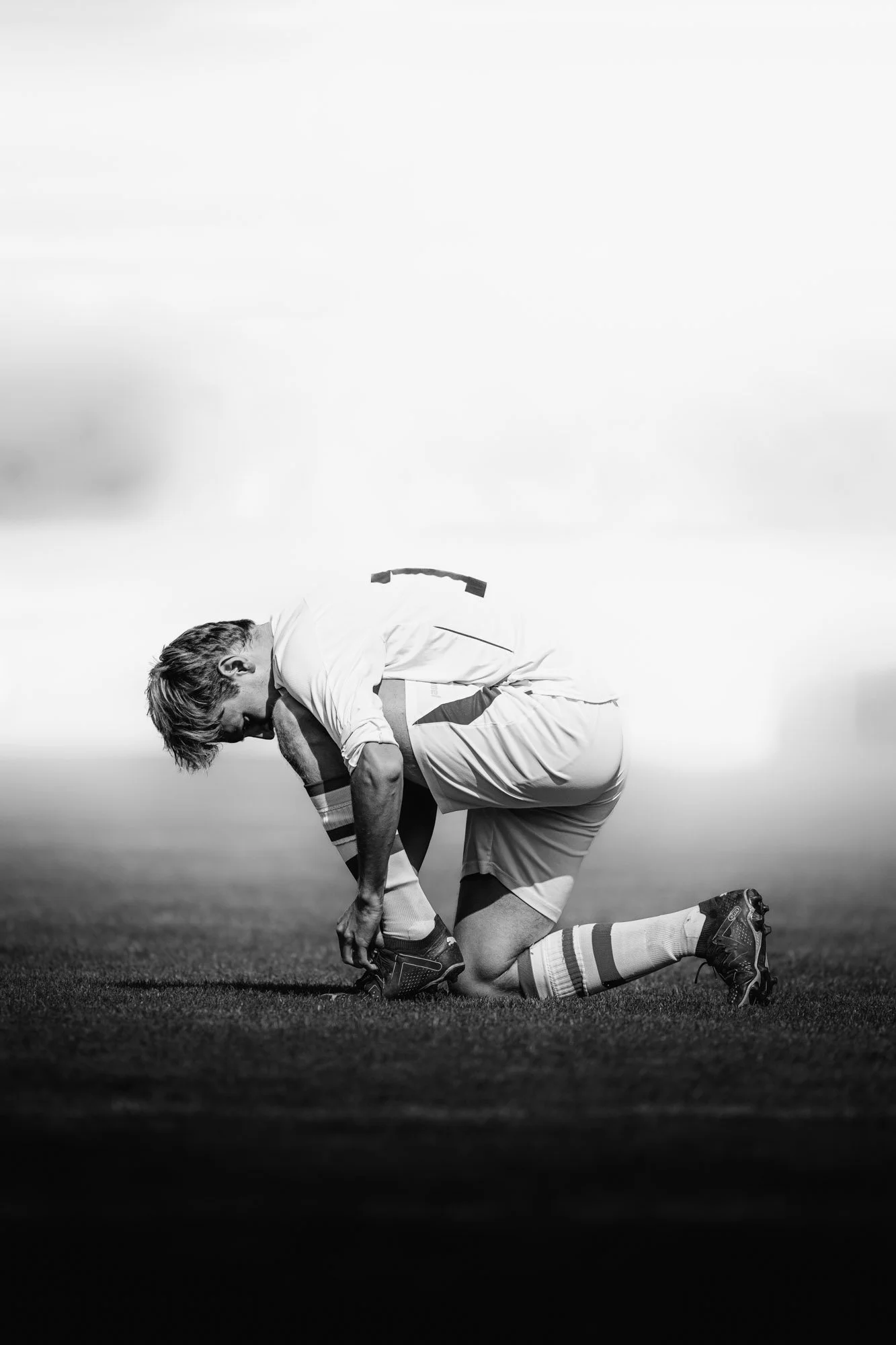 Een man in sportkleding knielt op een grasveld, boog voorover met beide handen op zijn schoenen tijdens zonsondergang.