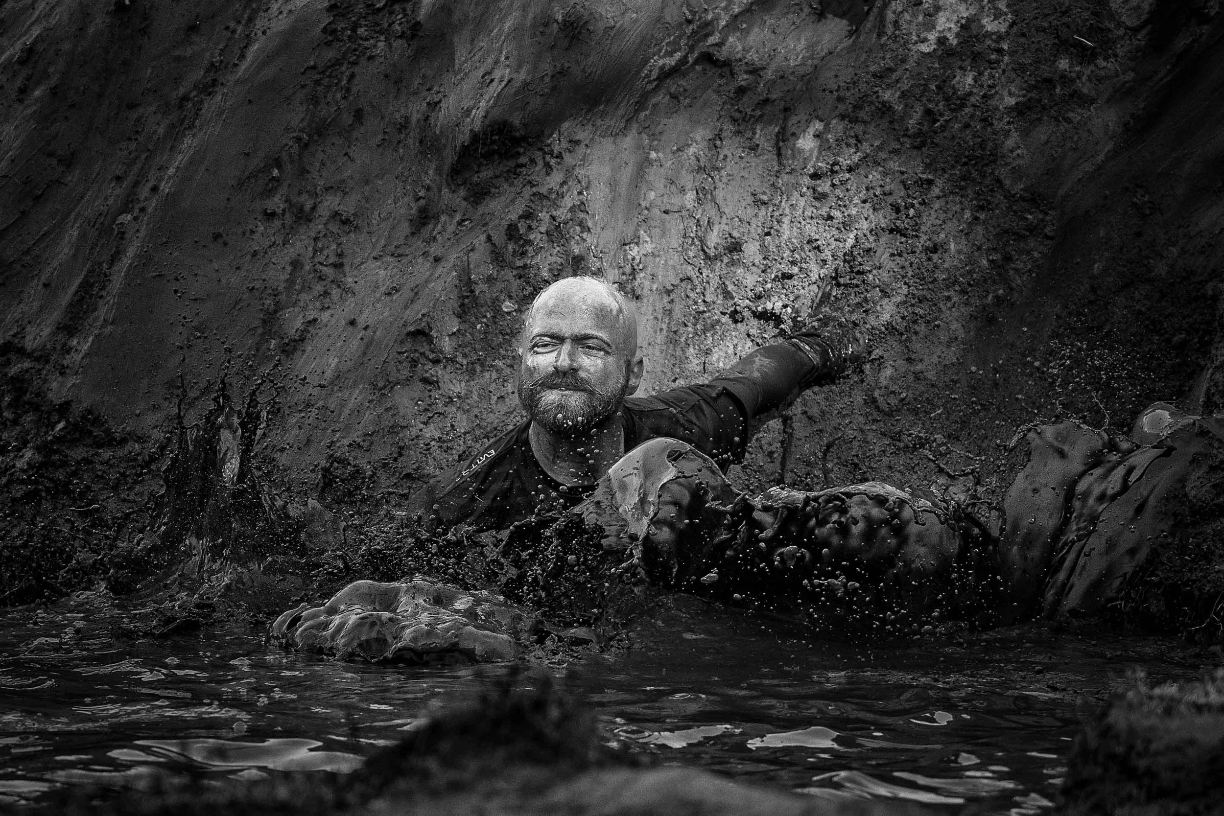 Man ligt in modder met water, glimlacht en kijkt omhoog, omringd door donkere aarde en modder.