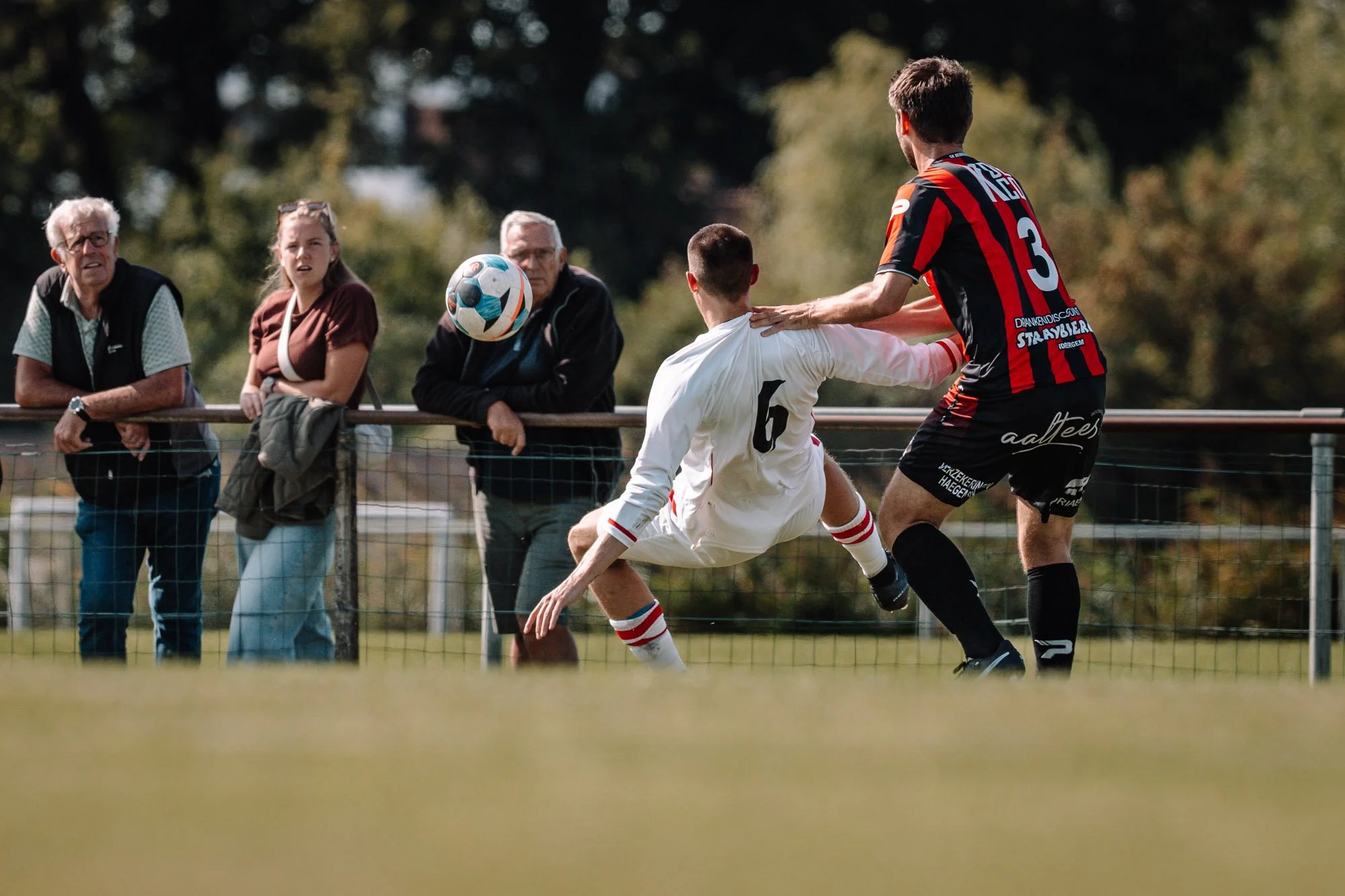 Twee mannen spelen voetbal op een veld terwijl een groep toeschouwers langs de zijlijn toekijkt.