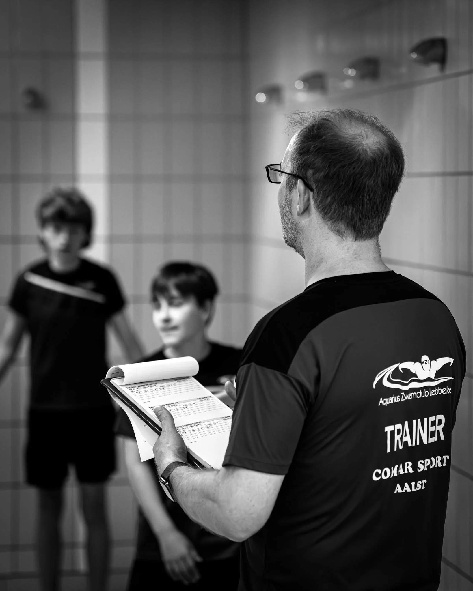 Een man met brillen en een zwart T-shirt dat 'Trainer COKAR Sport Aals' zegt, houdt een clipboard met papiertjes vast, terwijl hij in een sportschool of zwembadruimte staat. Op de achtergrond zijn mensen zichtbaar, enkele vrouwen met zwart sportoutfit.