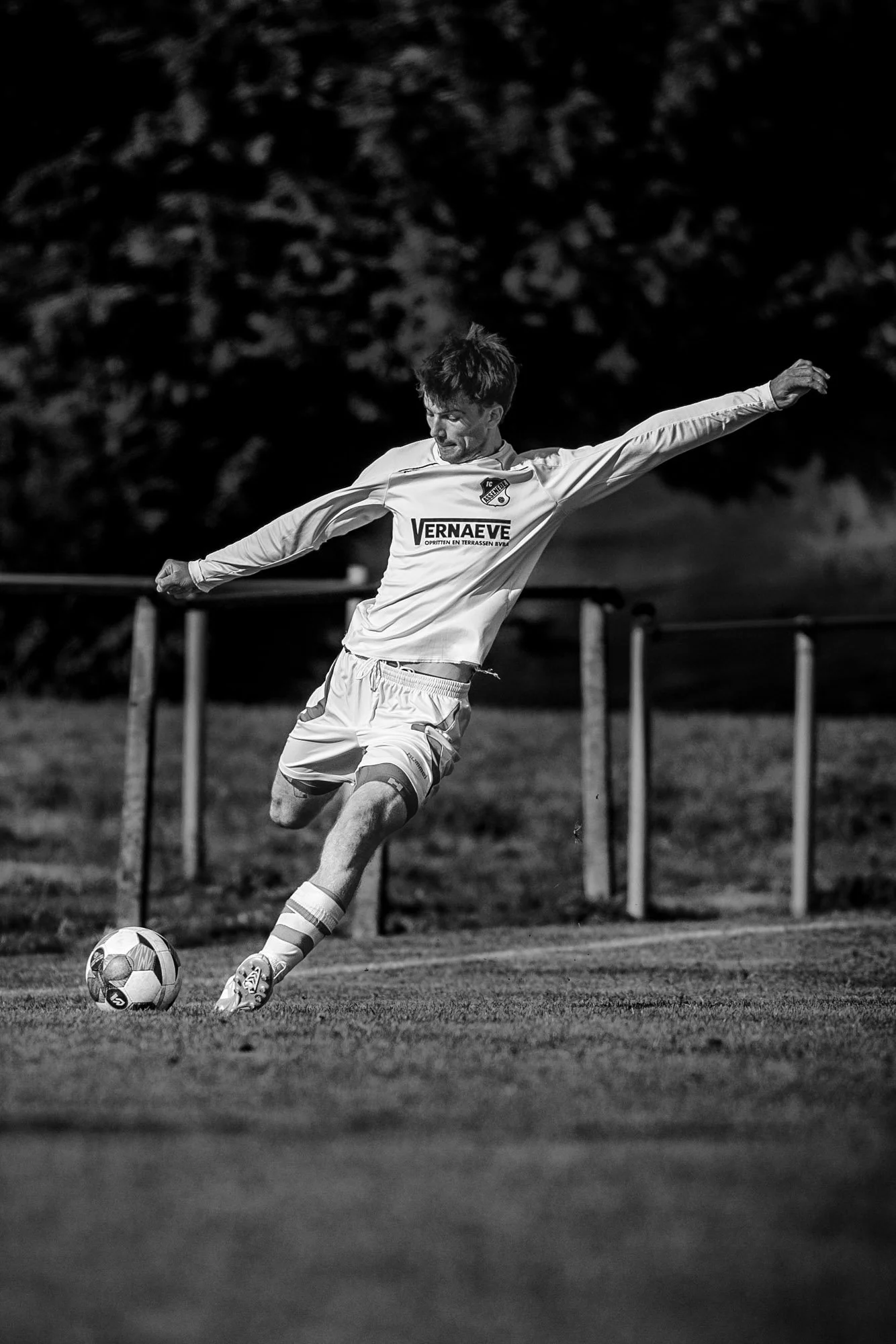 Zwart-witte foto van een man die een voetbal trapt op een grasveld, met een donkere achtergrond en schemerlicht.
