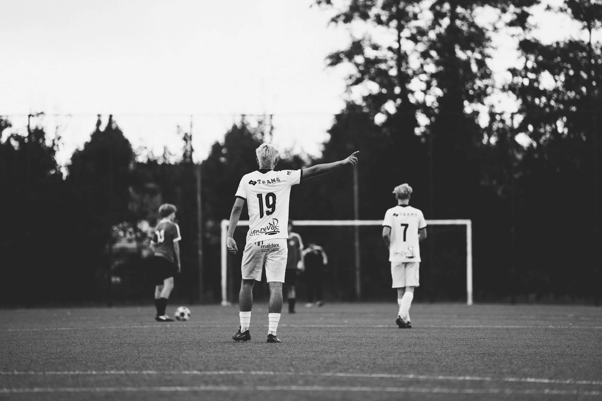 Zwart-wit foto van een voetbalwedstrijd met spelers op het veld, de speler met het nummer 19 wijst met zijn arm uit terwijl hij naar de bal kijkt, een ander spelertje met nummer 7 staat verder weg.