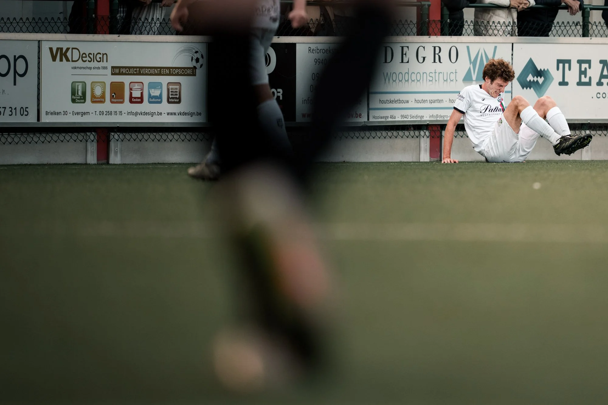 Een afbeelding van een voetbalwedstrijd met een speler die op de grond ligt terwijl hij een bal aan het ontglippen is. Op de achtergrond is een reclamebord te zien met verschillende advertenties.
