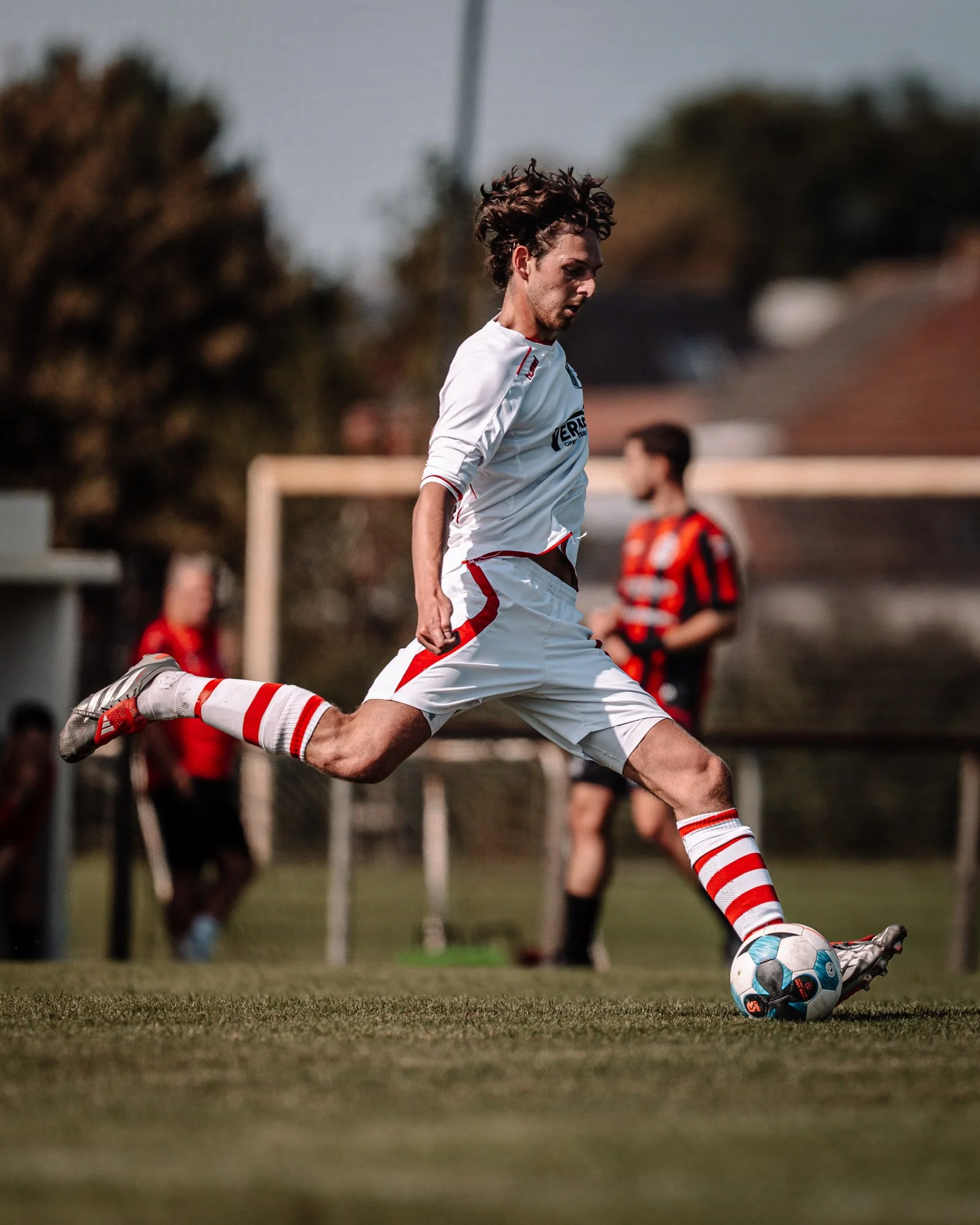 Voetballer in wit tenue trapt bal met rechtsvoet op grasveld, andere spelers op achtergrond, doelpost zichtbaar.