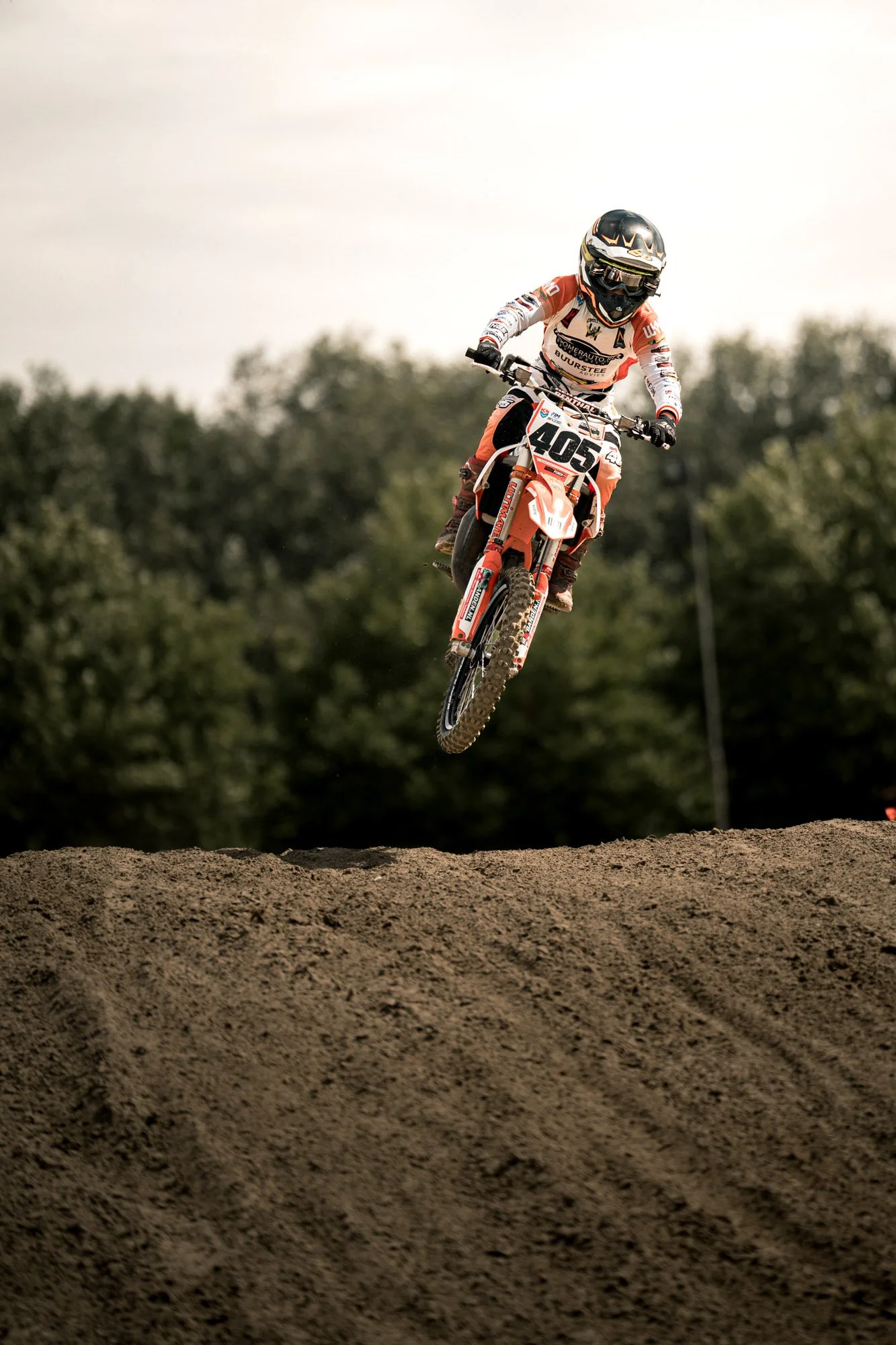 een motocrossracer die in de lucht springt op een dirttrack, met een achtergrond van bomen en een bewolkte hemel.