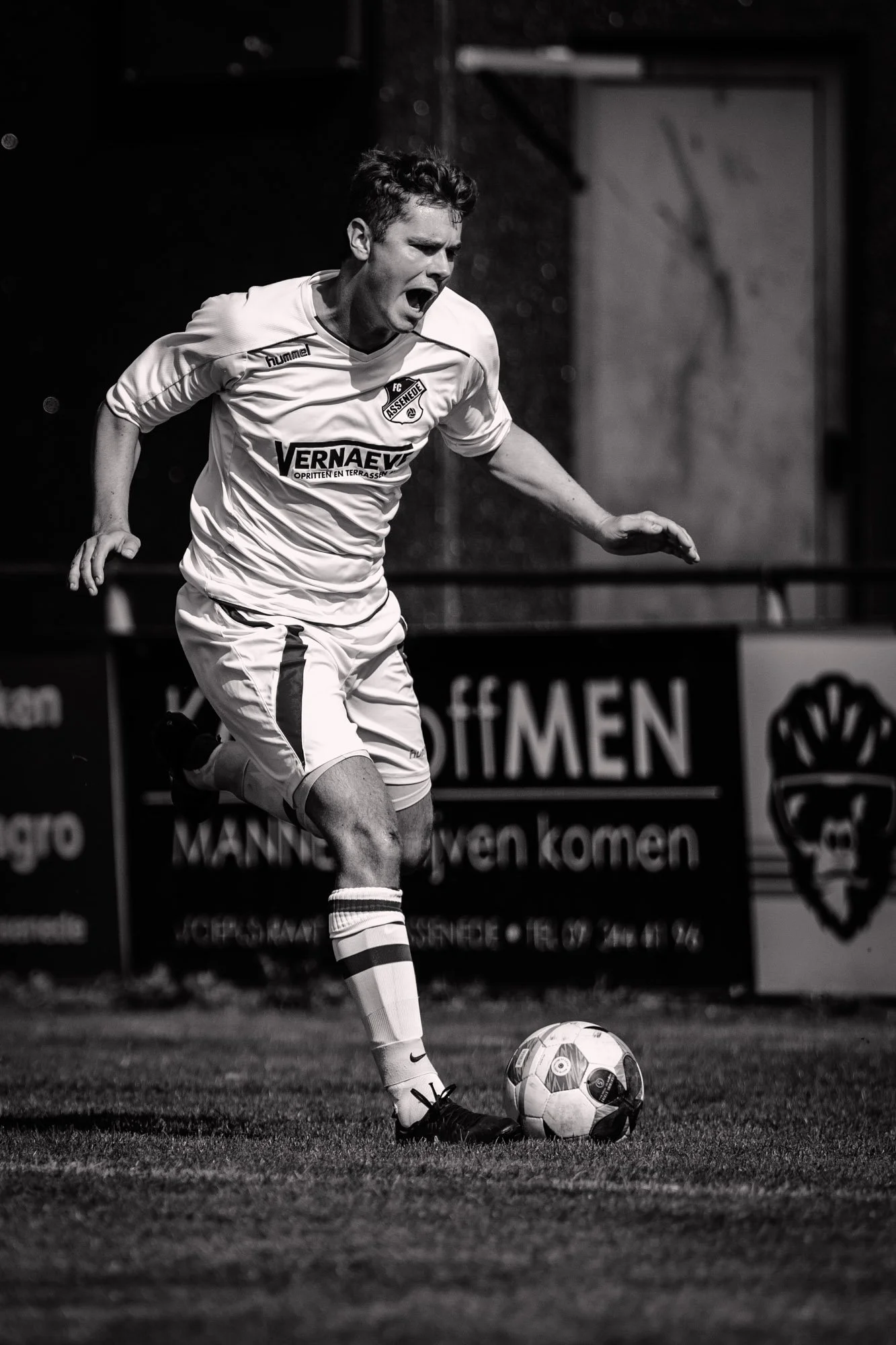 Zwart-wit foto van een man in een voetbaltenue die een voetbal trapt op een grasveld, met een muur en reclameborden op de achtergrond.