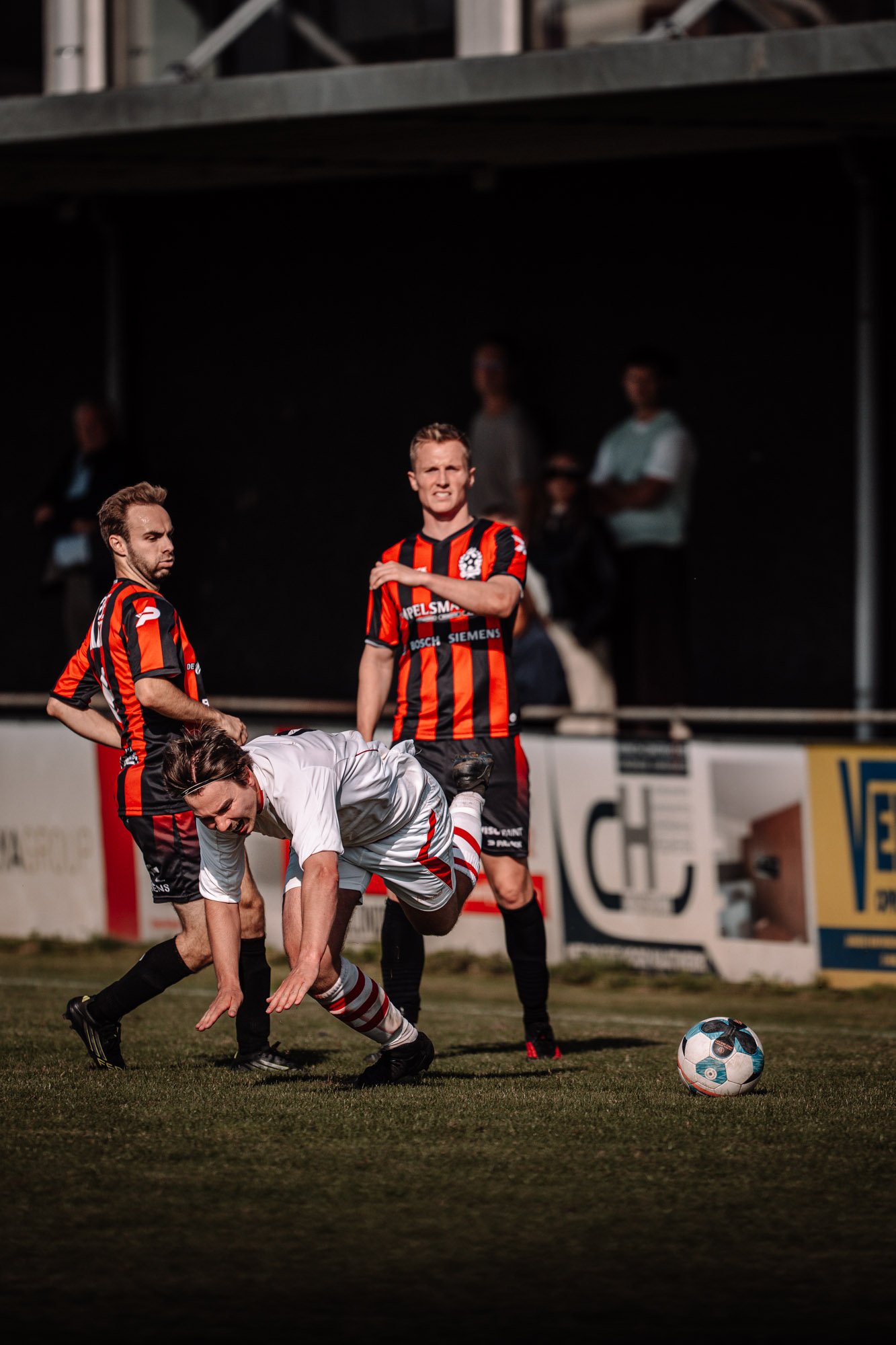 Een scène uit een voetbalwedstrijd waarbij spelers in rood-zwart gestreepte truien proberen de bal te bemachtigen, terwijl een speler in een witte uitrusting op de grond ligt en de bal voor zich heeft. Er zijn ook mensen op de achtergrond die naar de