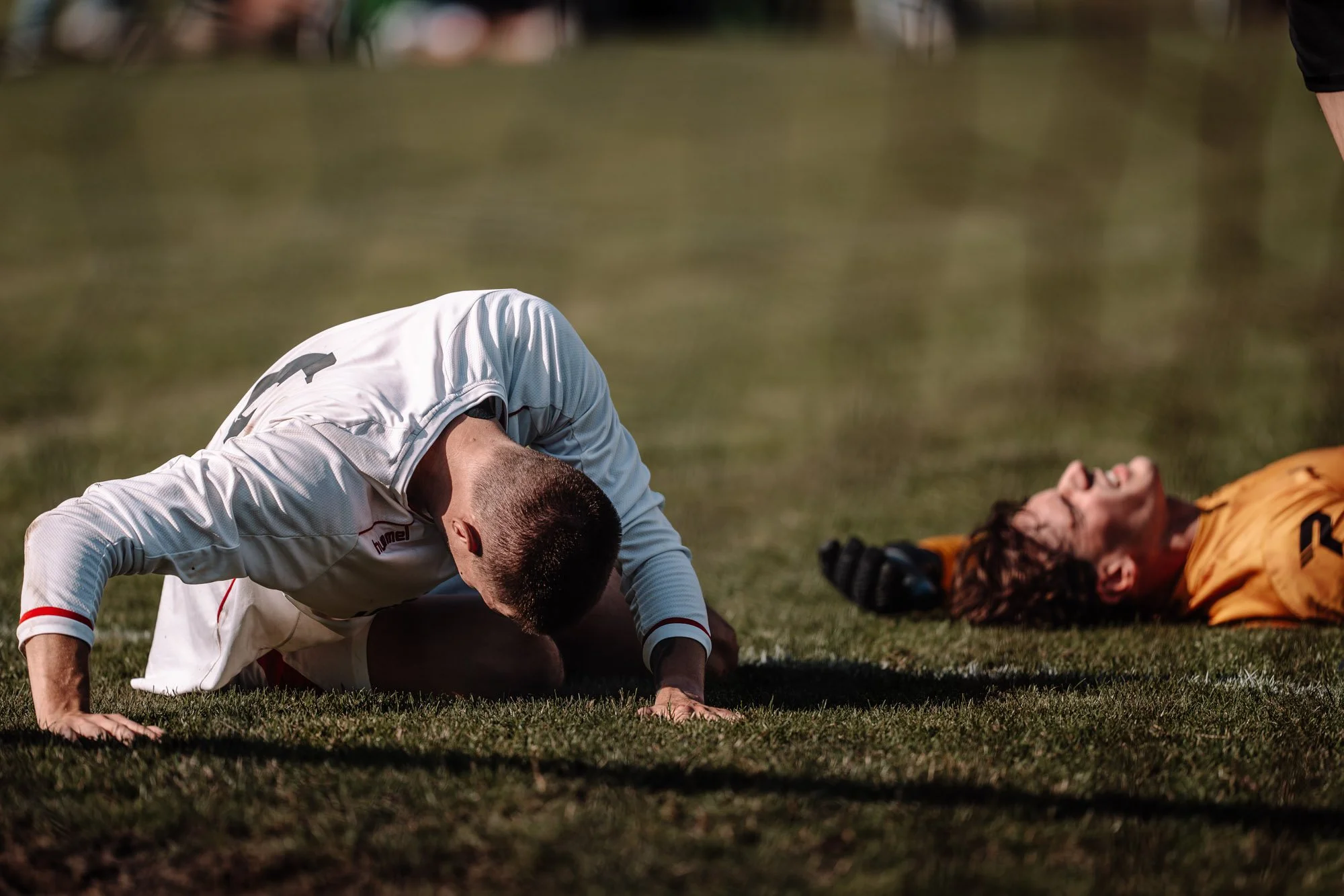 Een footballer ligt op de grond en lijkt bewusteloos, terwijl een ander speler en een persoon op de grond zich bezighouden met medische hulp op het veld.