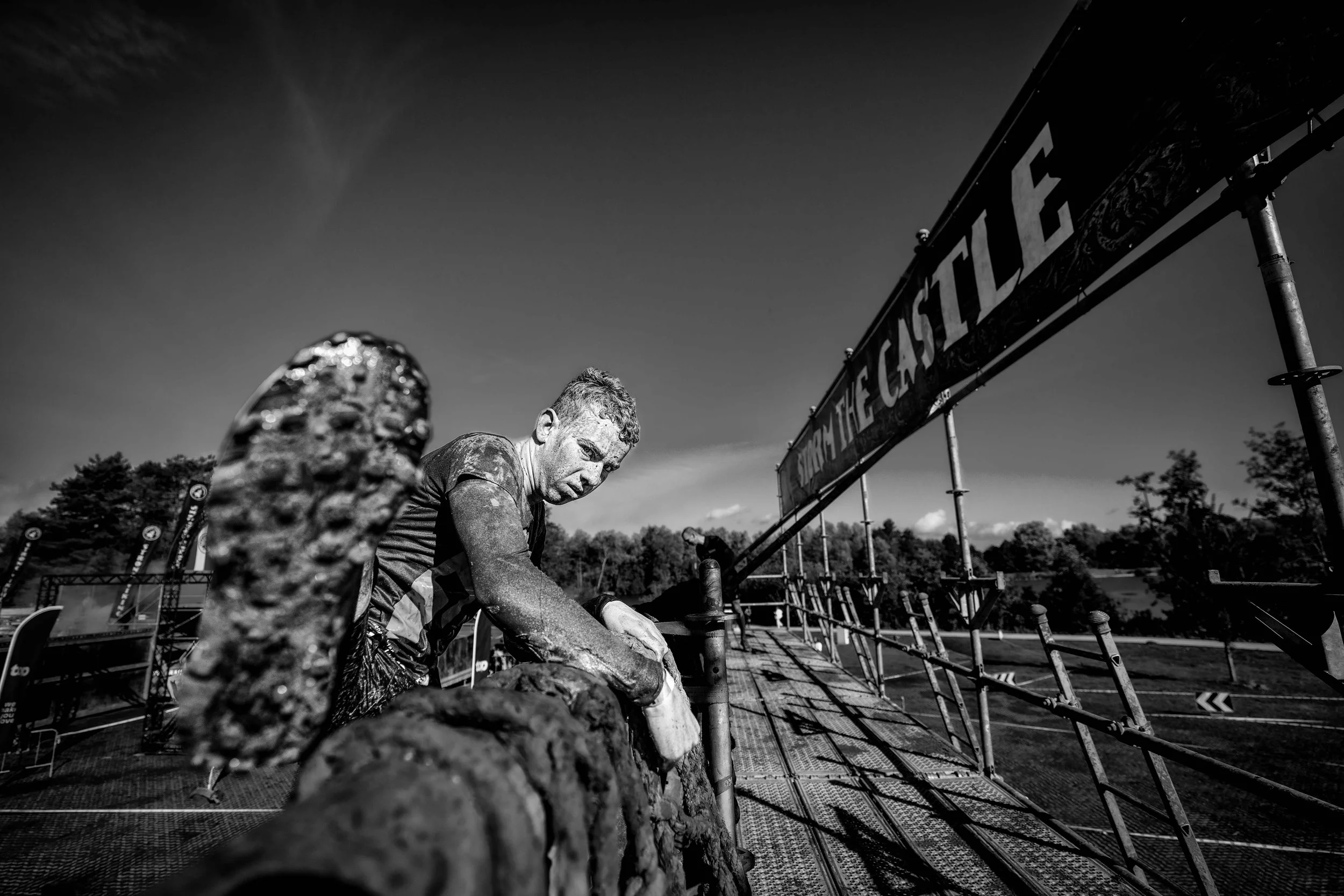 Een man die een obstacle race voltooit, bedekt met modder, onder een banner die zegt 'The Challenge', met een achtergrond van bomen en een blauwe lucht.