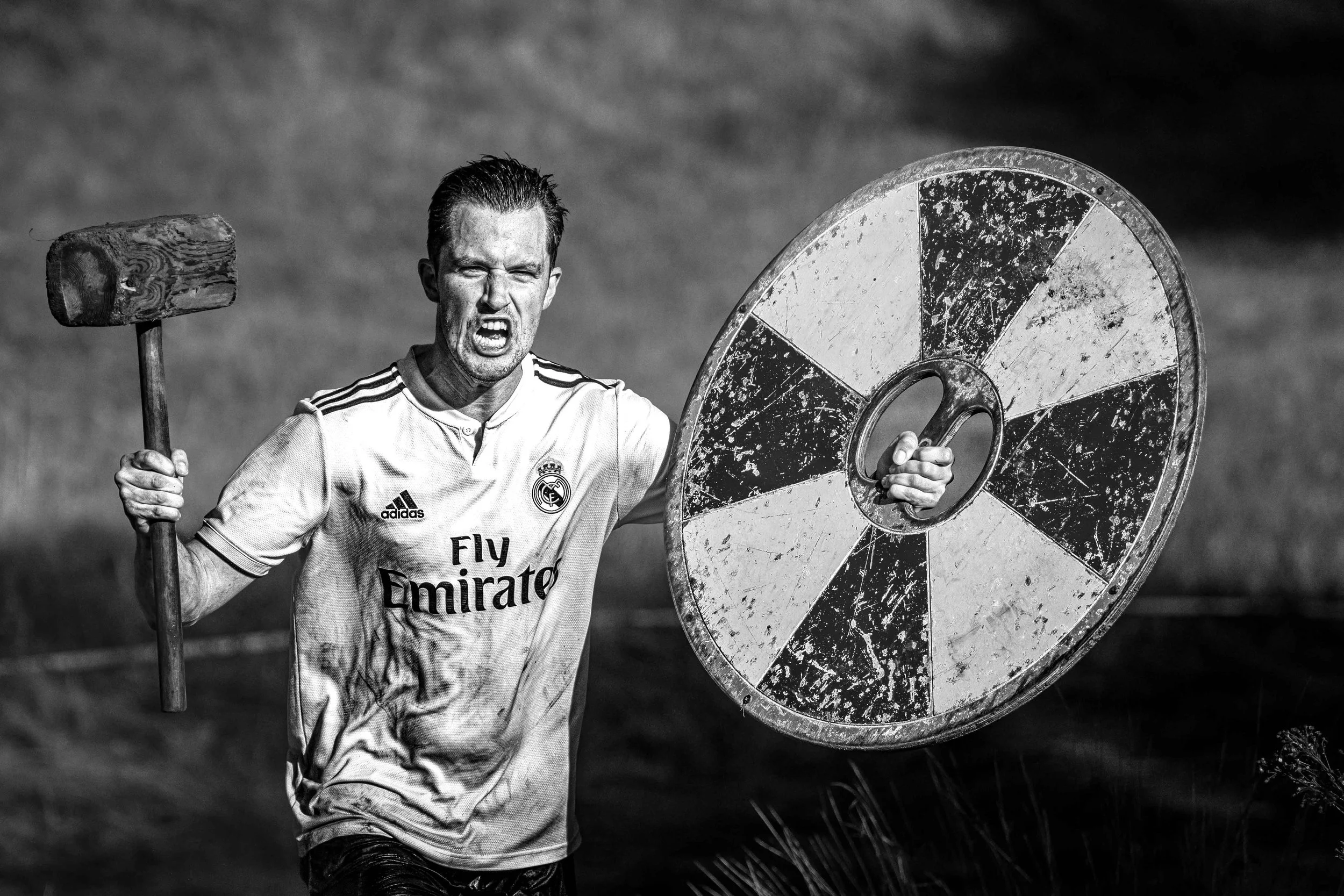 Man in een wit sportshirt met Real Madrid logo zwaait met een grote schijf en een hamer in een open landschap.