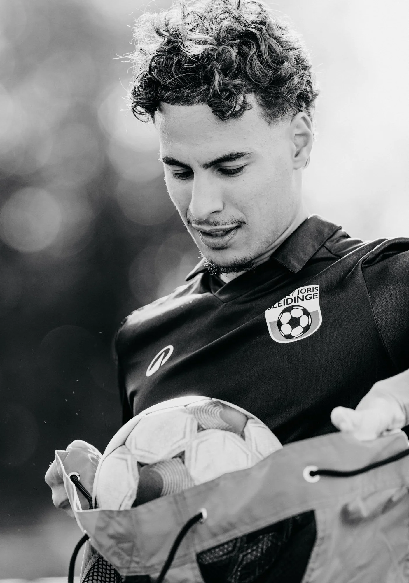 Een jonge man met krullend haar en een sporttenue die een bal vasthoudt, in een zwart-wit foto.