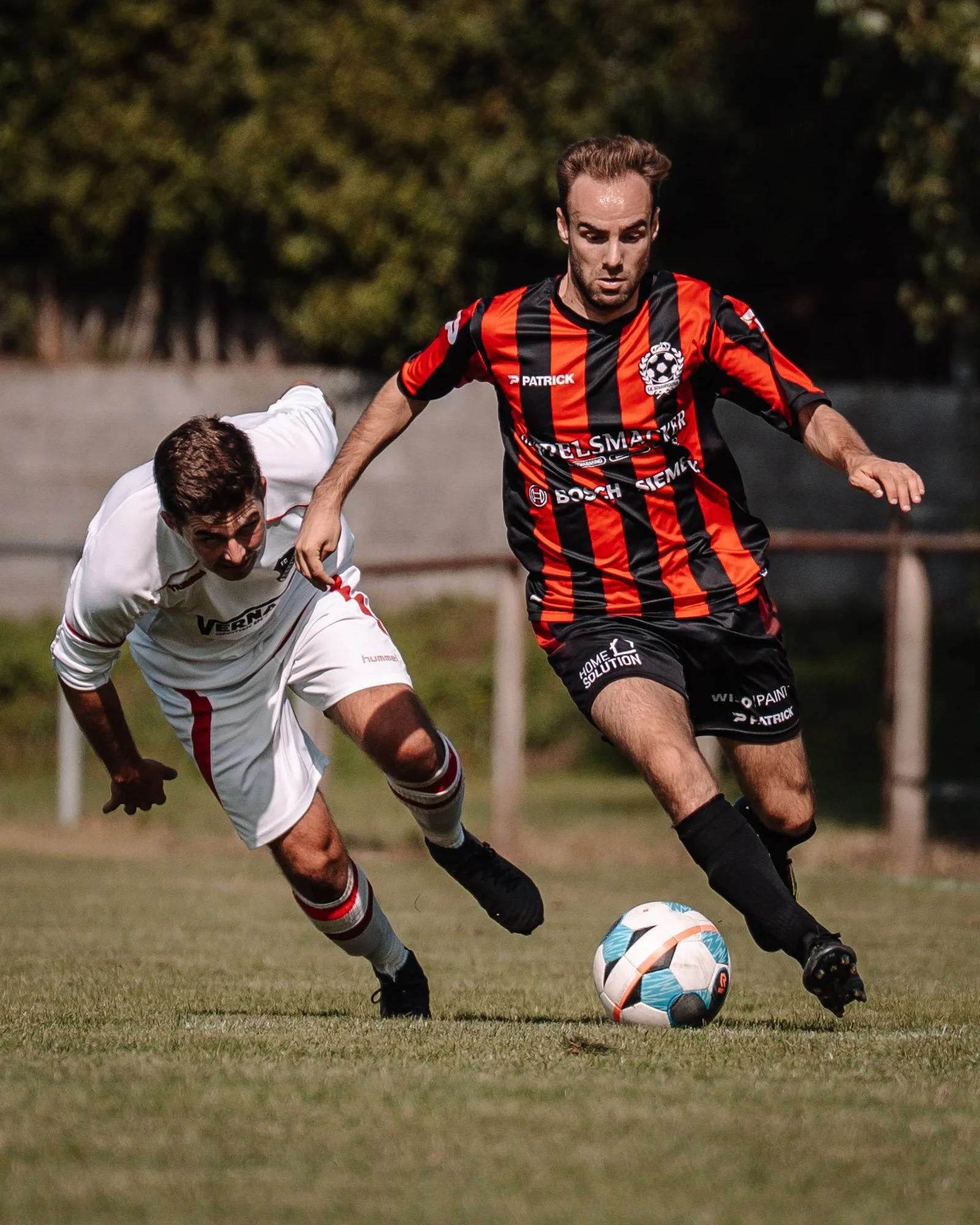 Twee voetballers in competitieve actie op het veld, met één die een bal probeert te controleren terwijl de ander probeert te blokkeren, omgeven door groene grasvelden en een bomenrij op de achtergrond.