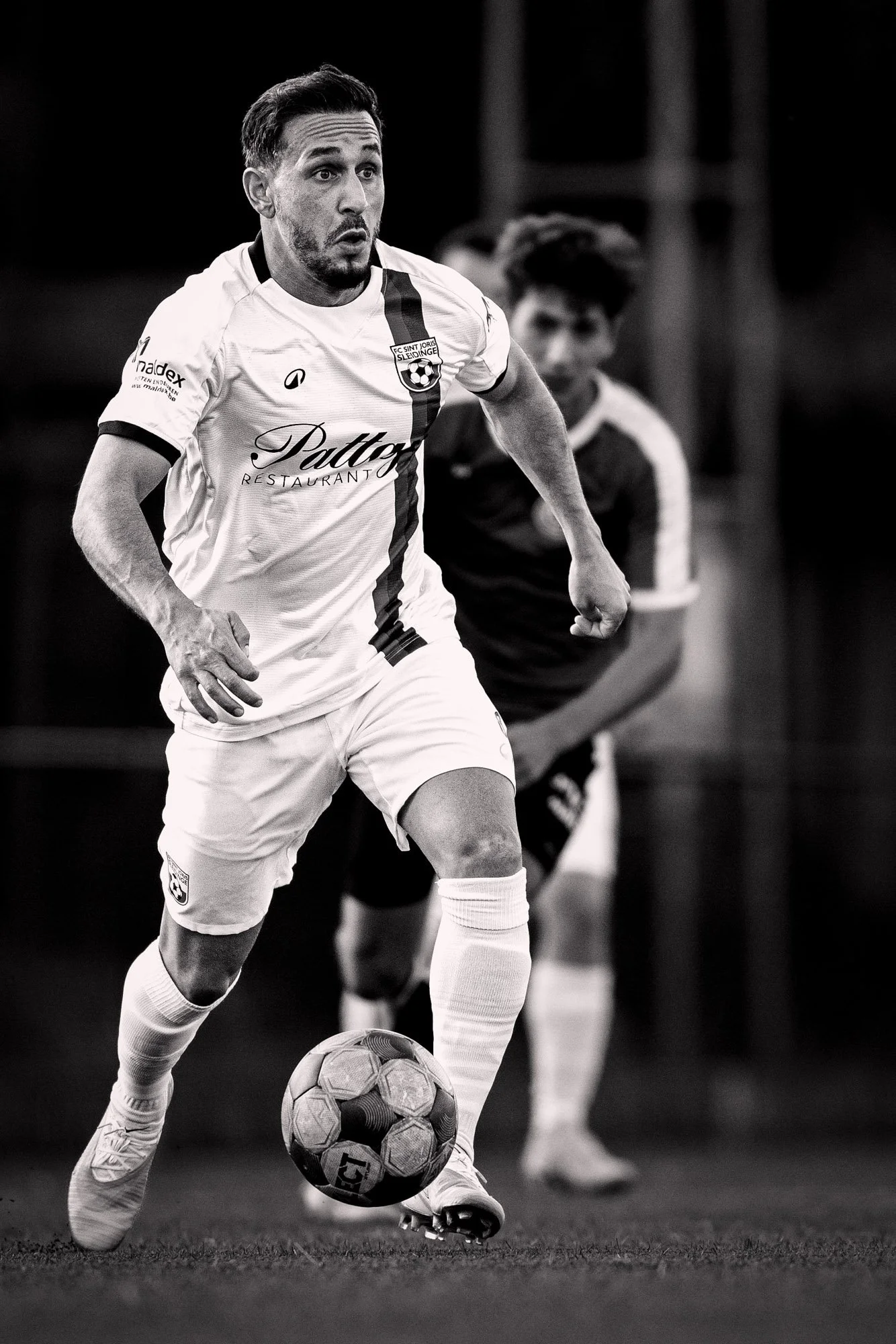 Zwart-wit foto van een man in een voetbalshirt die een wedstrijd speelt, met de bal onder zijn voet, op een grasveld.