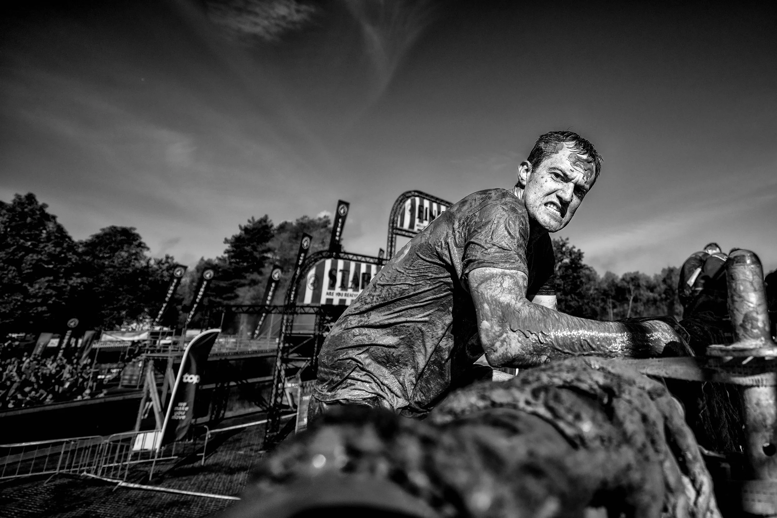 Man zit in modder, duidelijk uitgeput na een sportieve activiteit, met een achtergrond van een festivalterrein met borden en bomen, onder een bewolkte hemel.