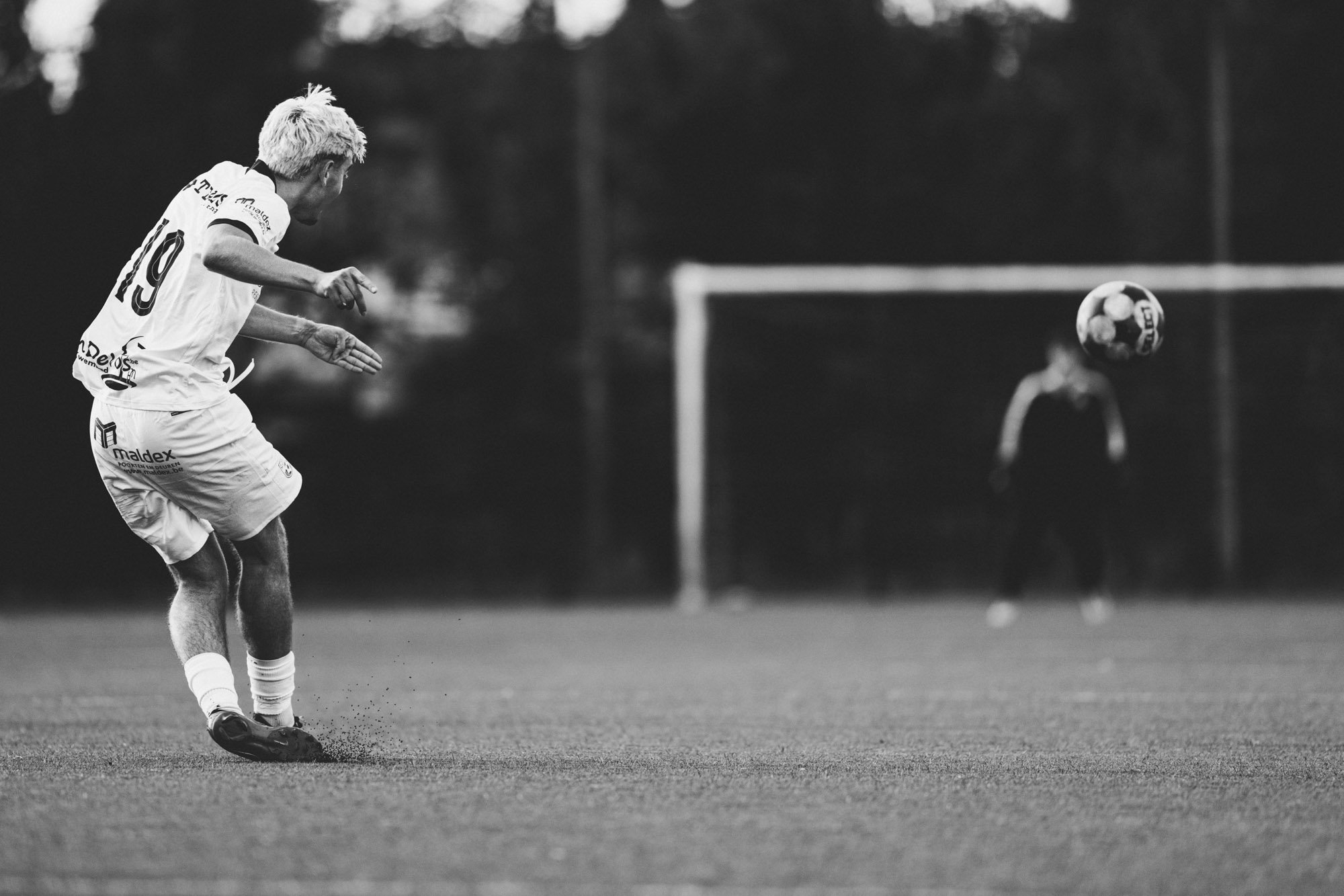 Zwart-wit foto van een man die net een bal trapt op een voetbalveld, met een doel en een speler op de achtergrond.