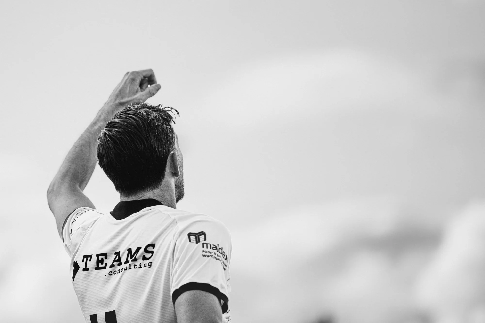 Zwart-witte foto van een man met kort haar en een sportshirt, gezien van achter. Hij heft één arm omhoog alsof hij de vuist in de lucht steekt. Op de achterkant van zijn shirt staat 'TEAMS . consulting'.