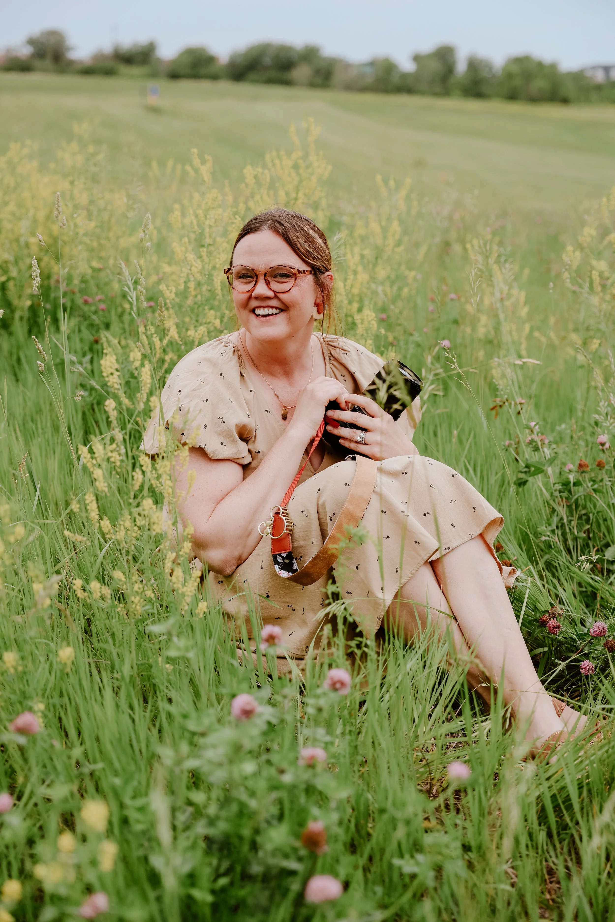A smiling woman with glasses and earrings sitting in a green field with yellow and pink flowers, holding a camera, during daytime.