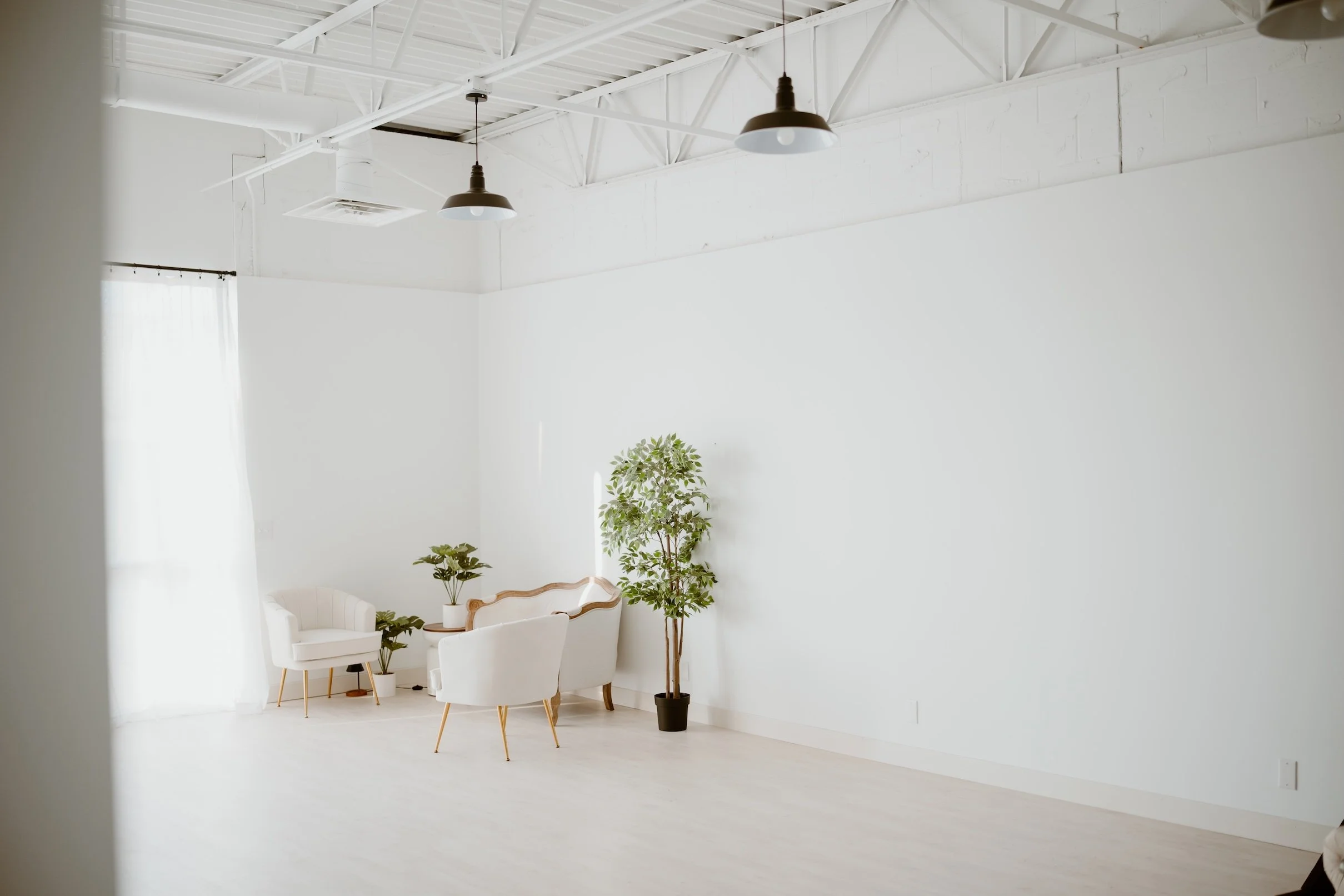 A bright, minimalist room with white walls, ceiling, and furniture, featuring two white chairs, plants, and a large window with sheer curtains.