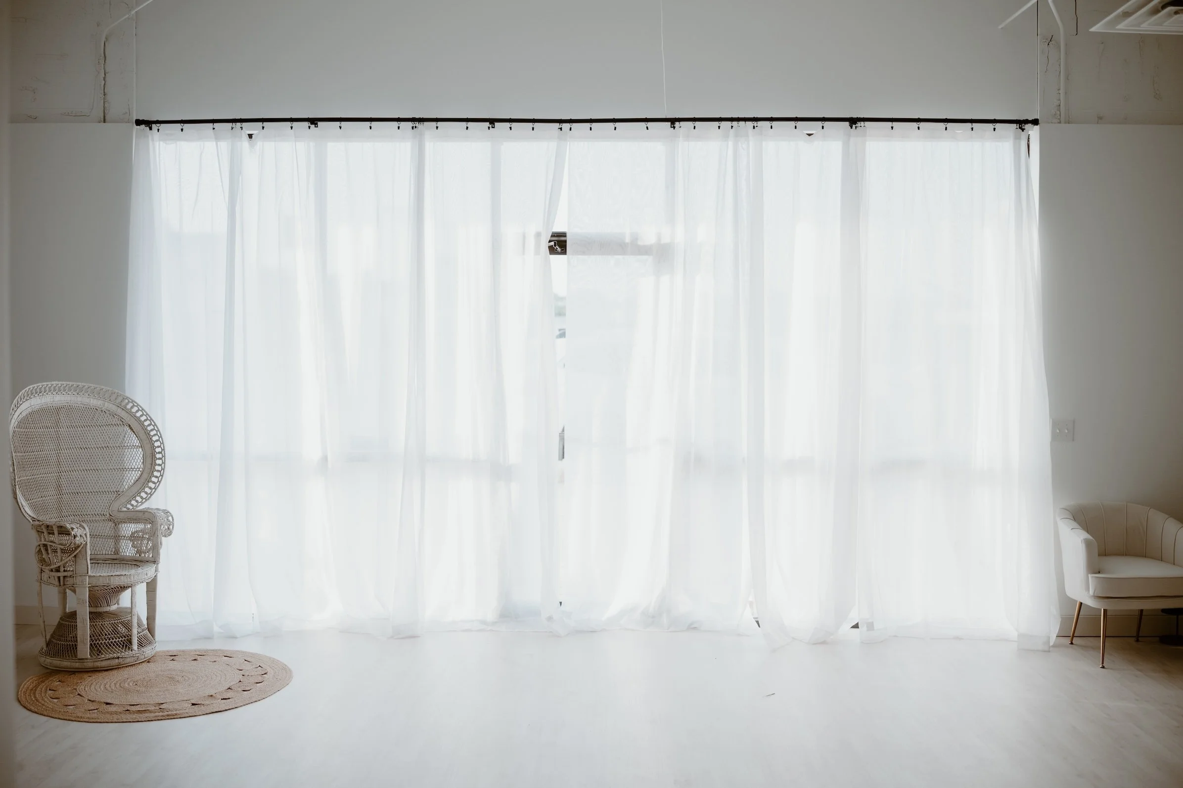 Bright room with sheer white curtains covering a large window, two woven chairs on the left, a small beige rug, and a white armchair on the right.