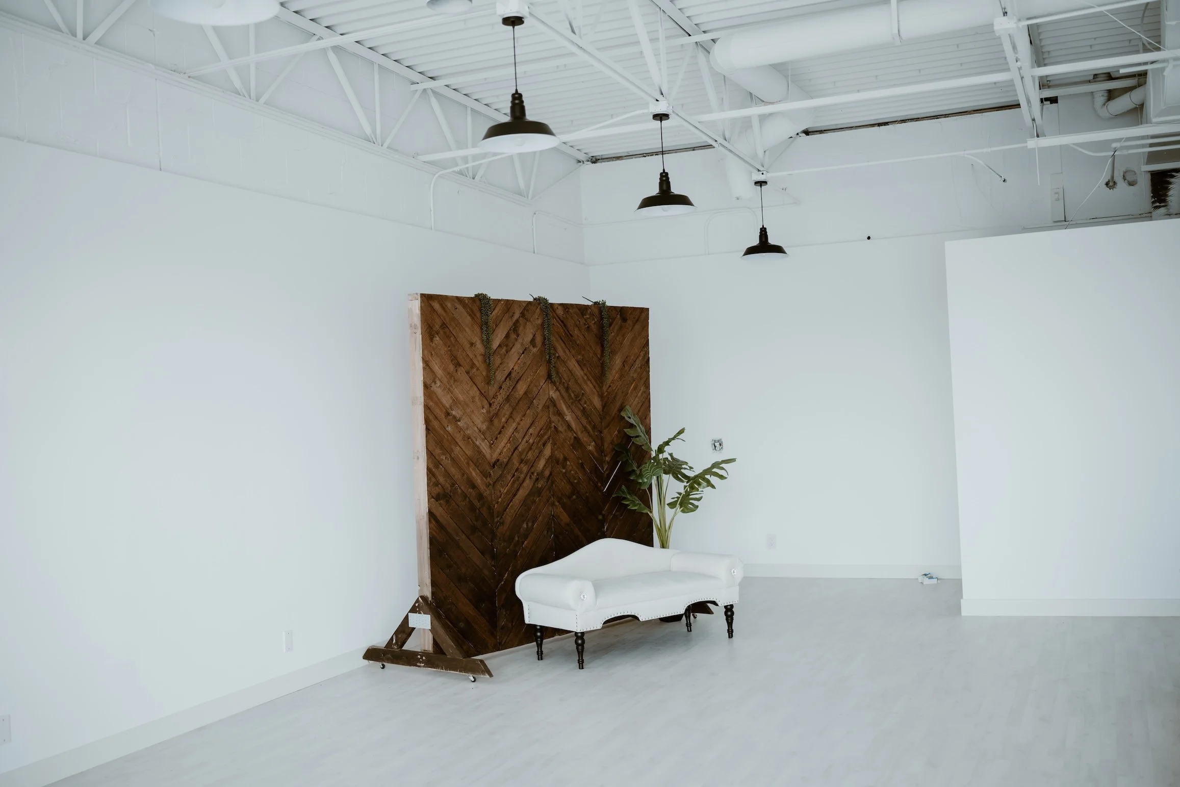 Interior of a modern industrial style space with white walls and ceiling, featuring black hanging pendant lights, a wooden partition, a white tufted bench, and some green plants.