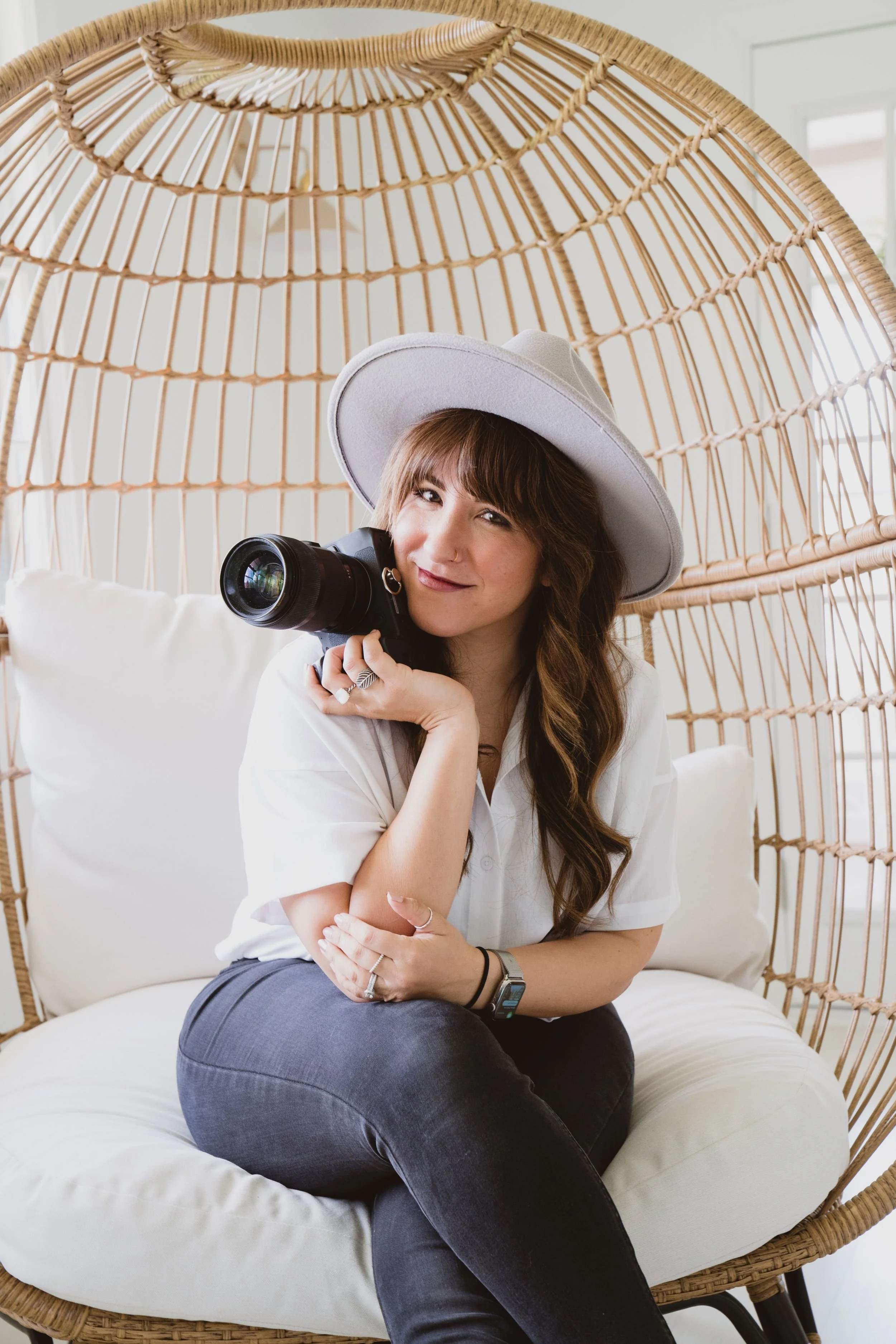 A woman with long brown hair, wearing a light gray hat, white shirt, and dark gray pants, sitting on a white cushioned chair inside a wicker hanging chair. She is holding a camera near her face, smiling slightly, with her arms crossed and in a relaxed pose in a bright room with white walls and a window in the background.