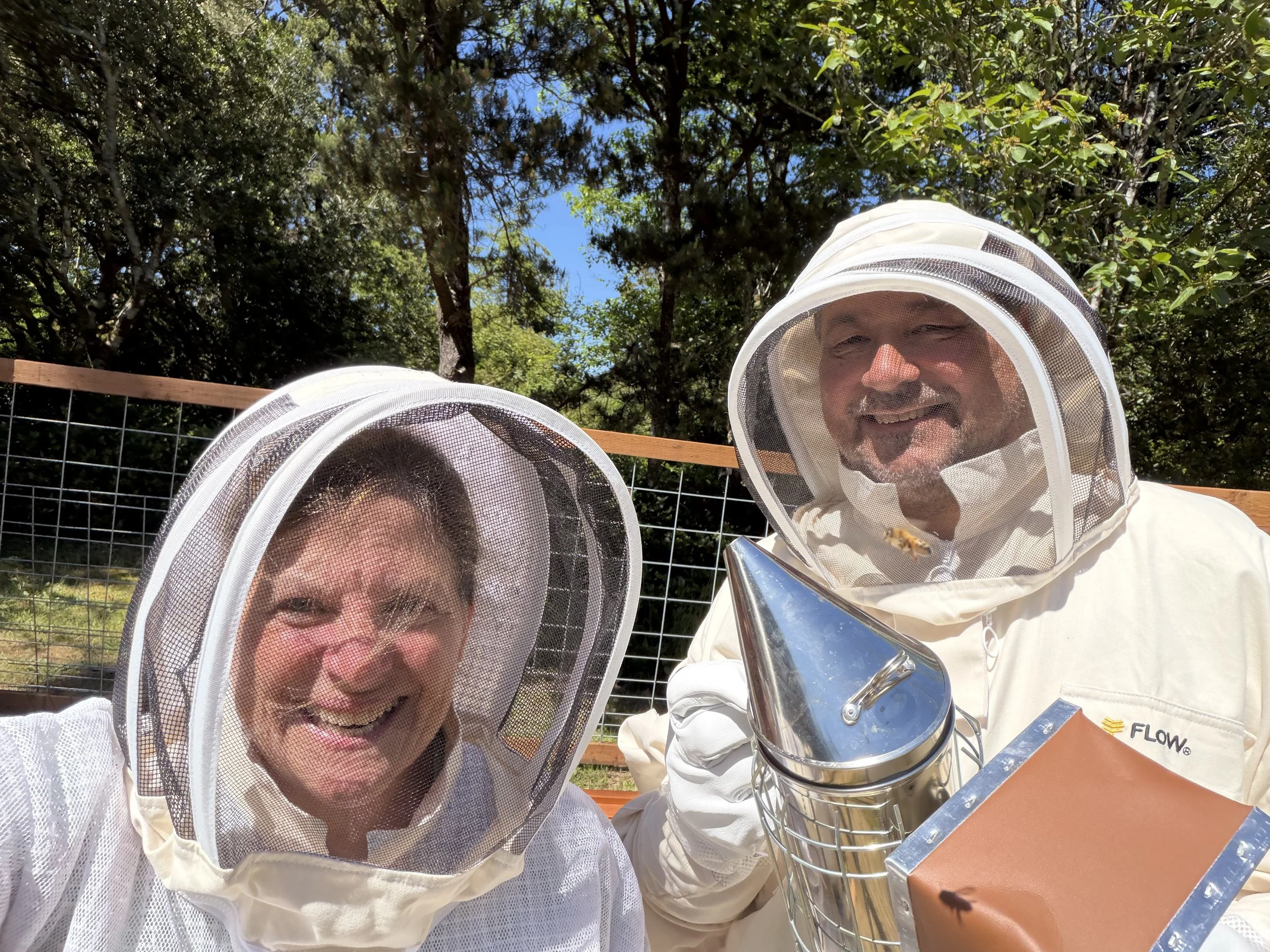 Two people in beekeeping suits and veiled hats are smiling outdoors, with trees and blue sky in the background. They are holding honey harvesting equipment, including a large metal container with bees on it.