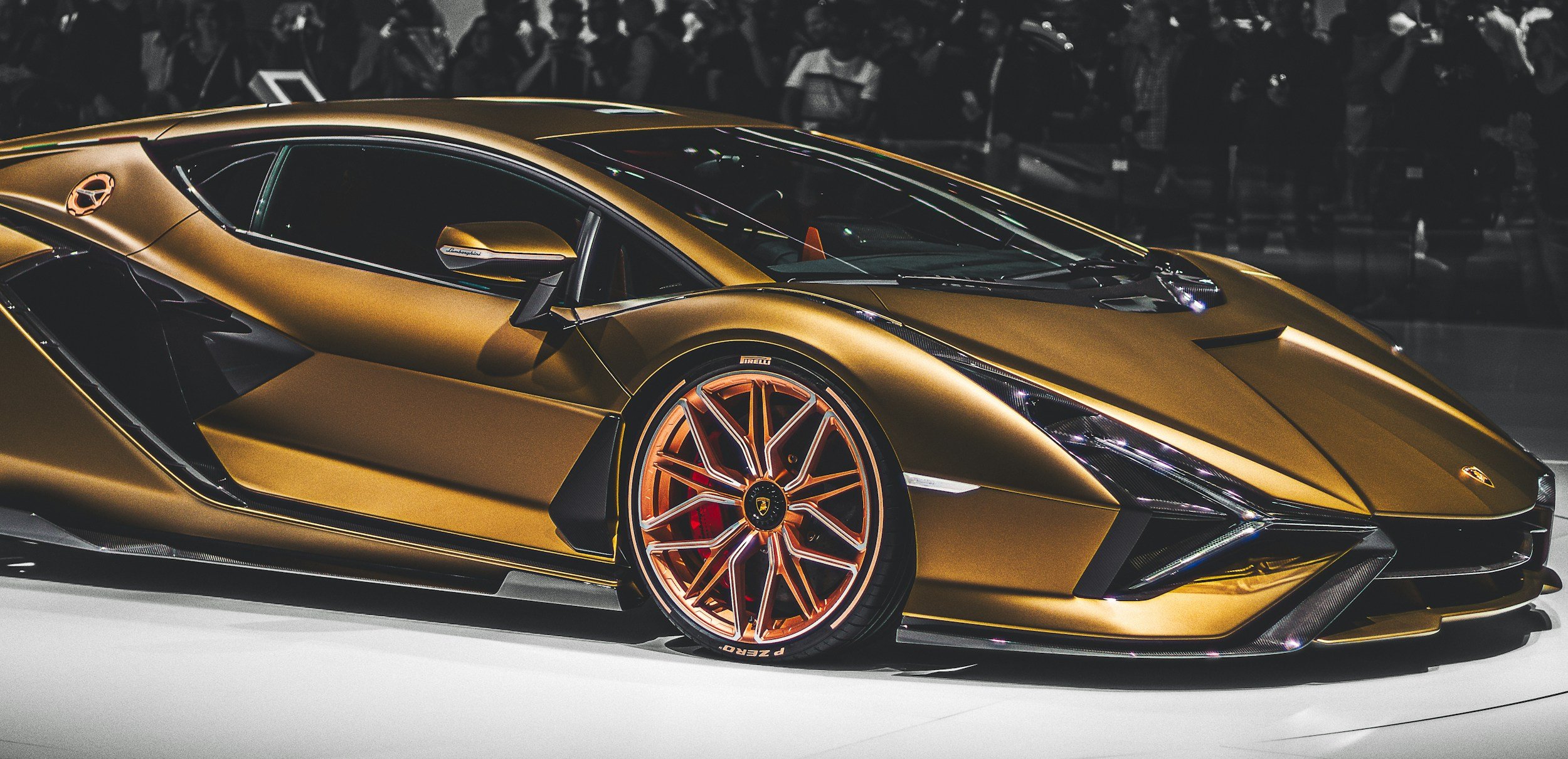 A gold Lamborghini sports car on display at an auto show, with a crowd of people in the background.