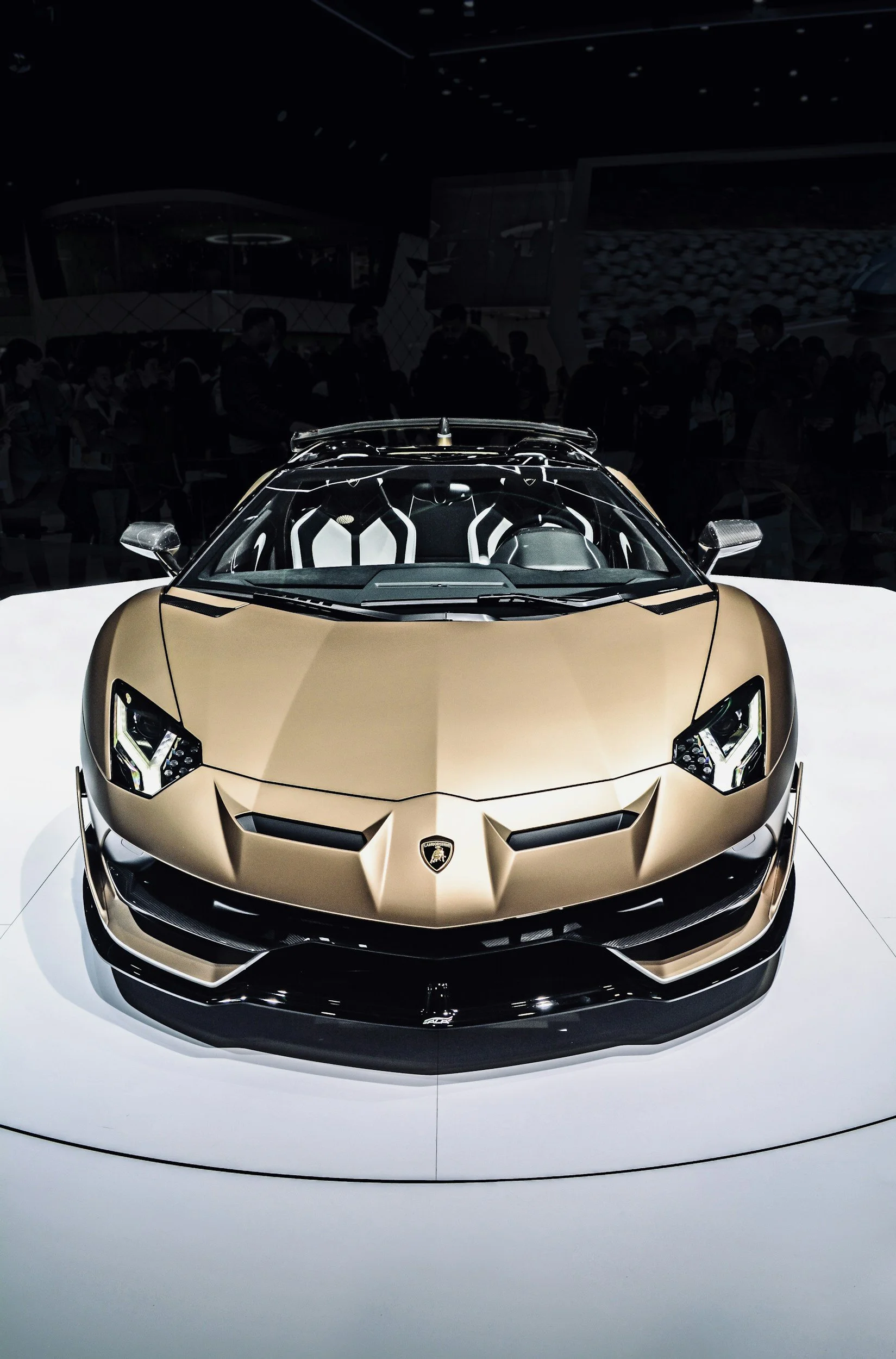 A gold-colored Lamborghini sports car displayed at an auto show, with people in the background observing the vehicle.