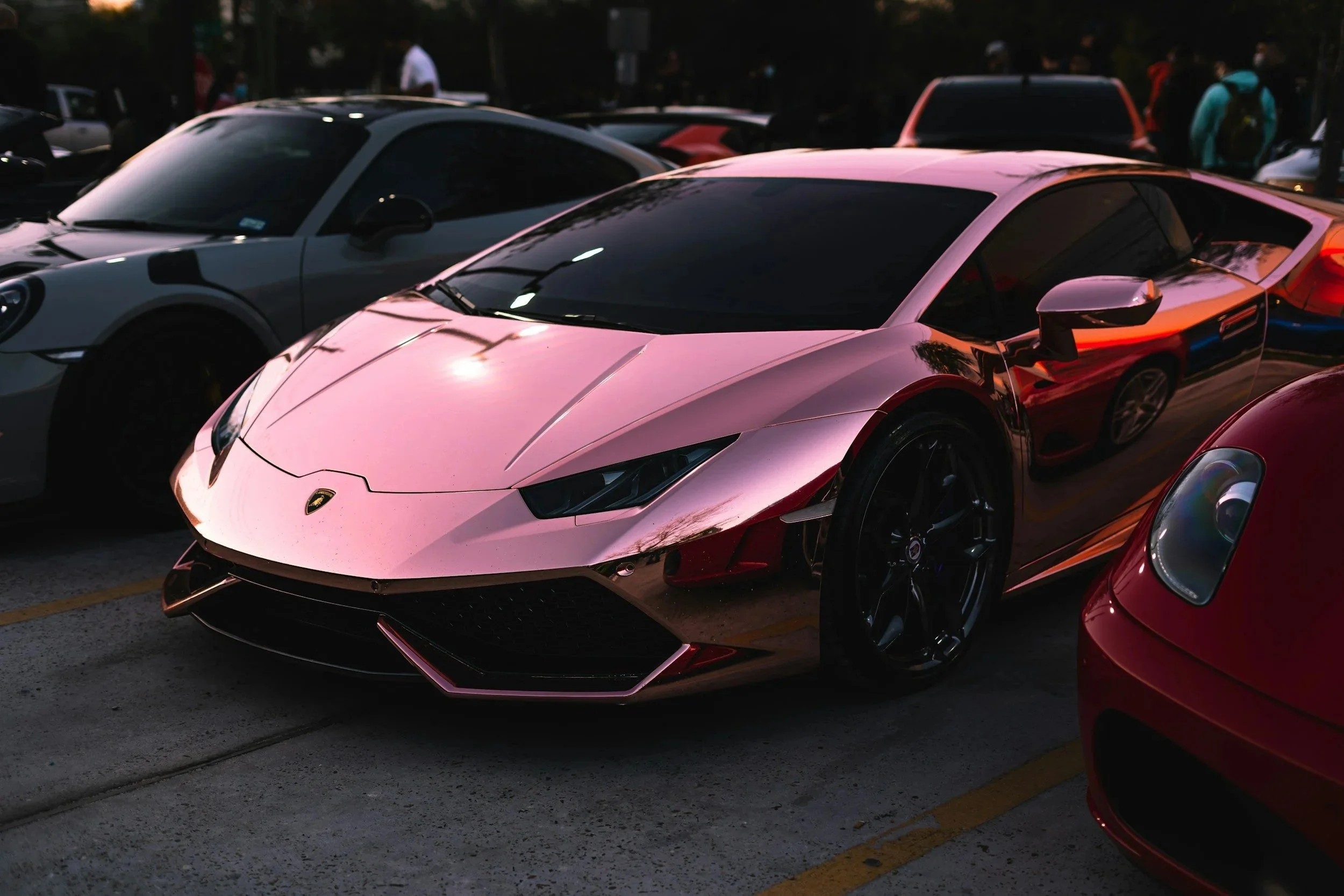 A pink Lamborghini sports car parked among other vehicles in a parking lot during dusk.