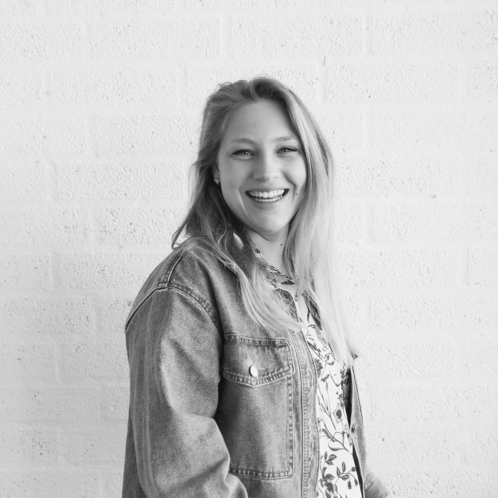 Zwart-wit foto van een lachende jonge vrouw met lang haar, gekleed in een denim jas, staand voor een witte muur.