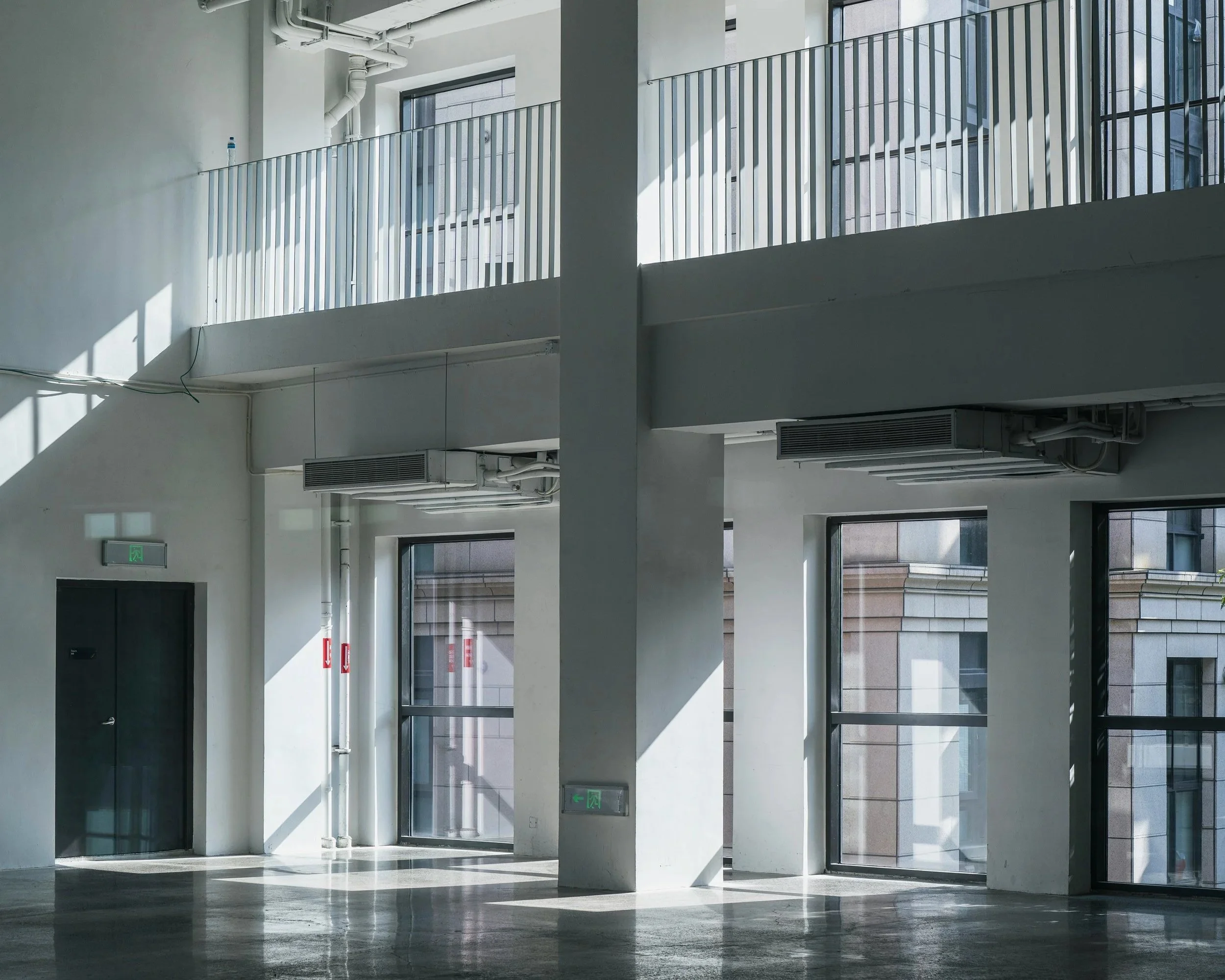 Interior de un edificio moderno con ventanas grandes, paredes blancas y luces naturales, incluyendo puertas de entrada y signos de salida de emergencia.