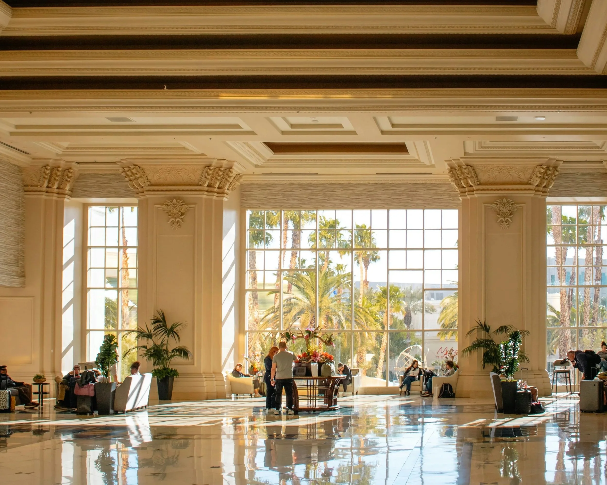 Lobby de hotel elegante con grandes ventanales, plantas y personas sentadas y conversando.