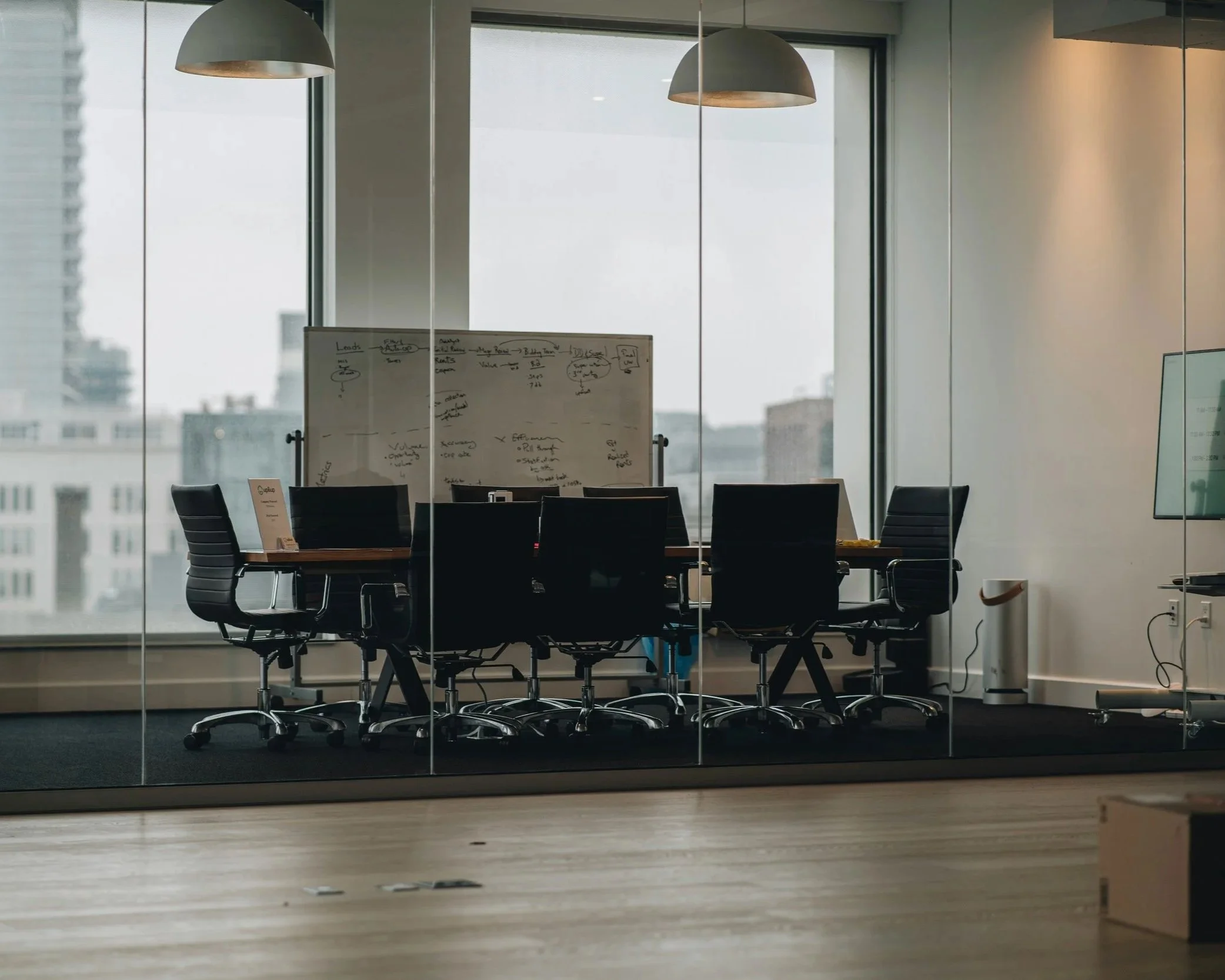 Sala de reuniones con mesa grande y sillas negras, vista a través de un vidrio con reflejo, en una oficina moderna con ventanas grandes que dejan entrar luz natural.