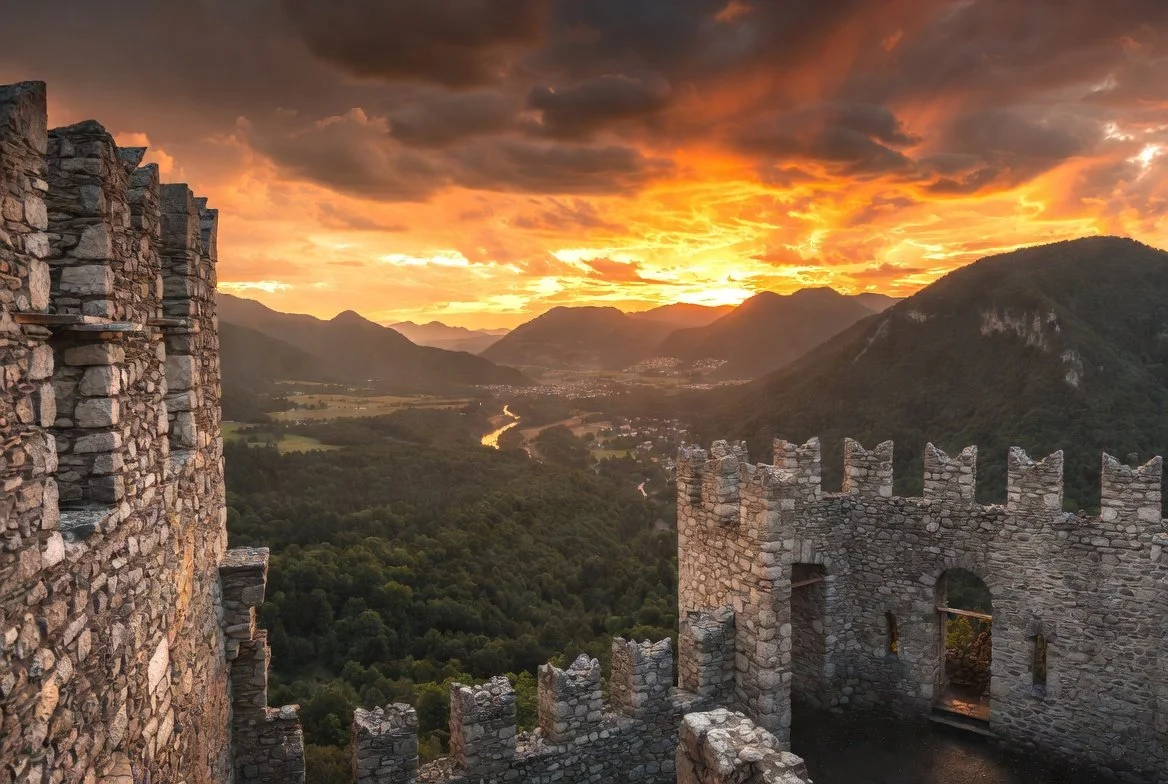 Stone castle walls at sunset with a valley, river, and mountains in the background.