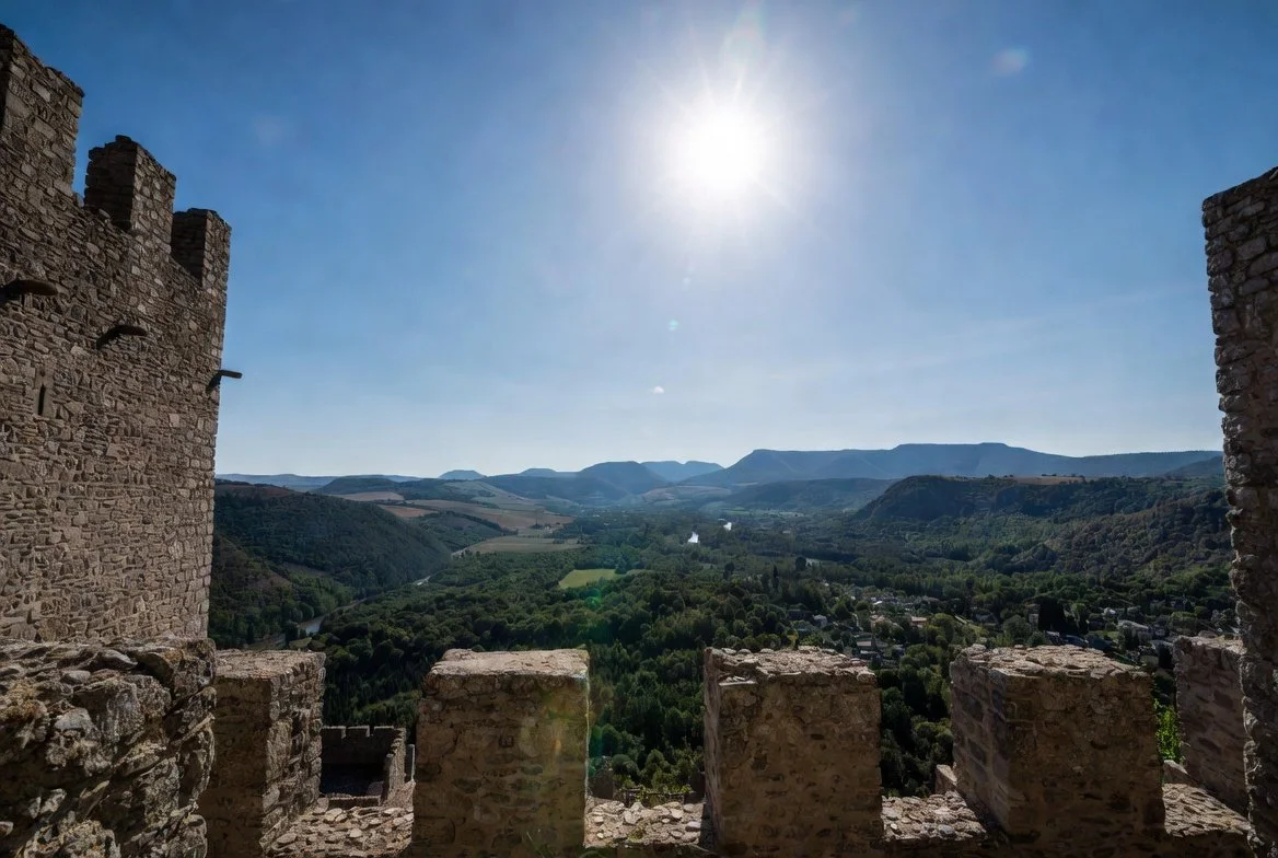 Stone castle ruins overlooking a lush green valley with mountains in the distance under a bright sun.