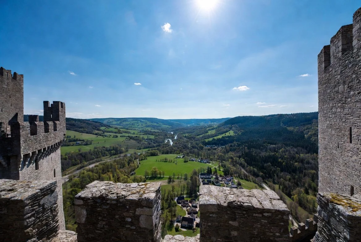 View from a castle tower overlooking a green valley with hills, trees, a river, and scattered houses under a blue sky with some clouds.