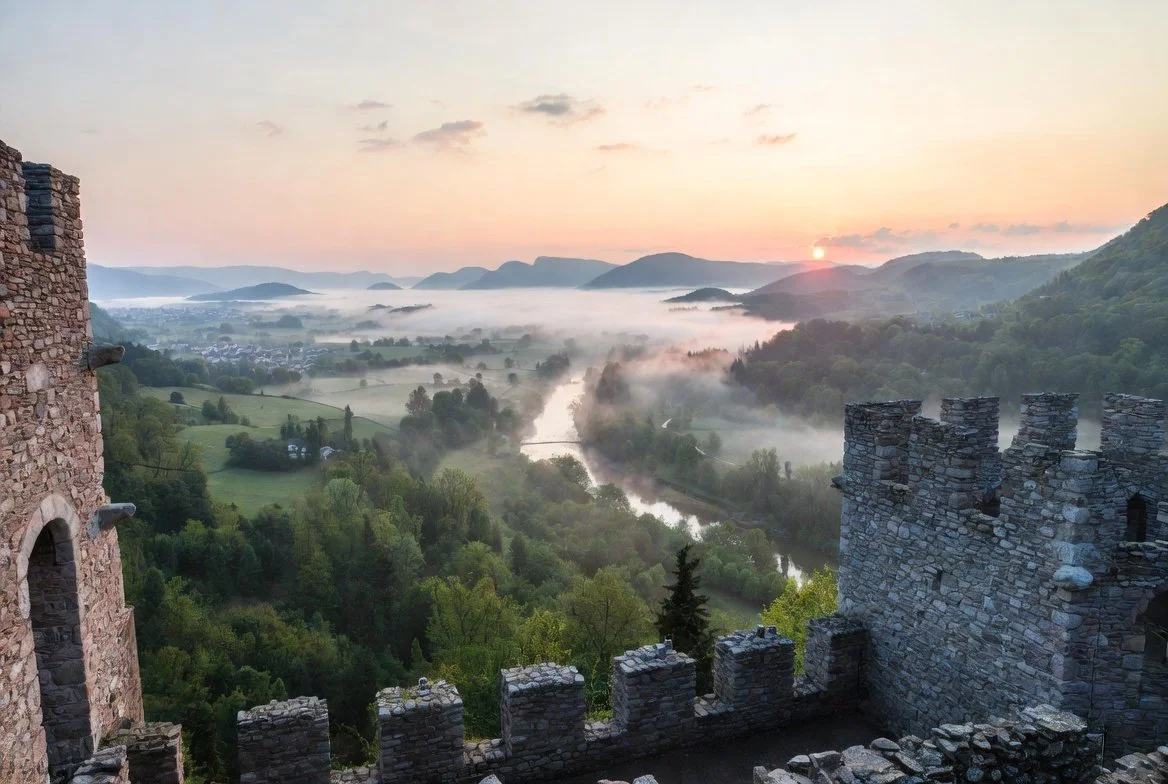 Sunrise over a lush green valley with a river, surrounded by hills and mountains, viewed from a stone castle with battlements.