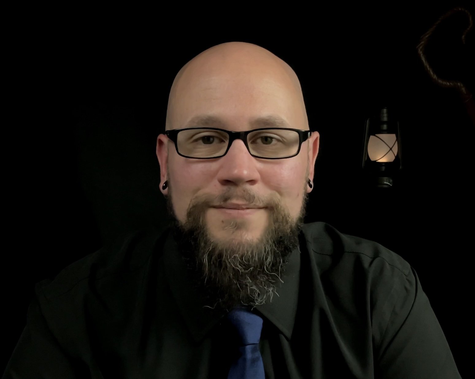 A man with a bald head, glasses, and a beard, smiling at the camera. He is wearing earrings and a black shirt with a blue tie. There is a lantern on the black background behind him.