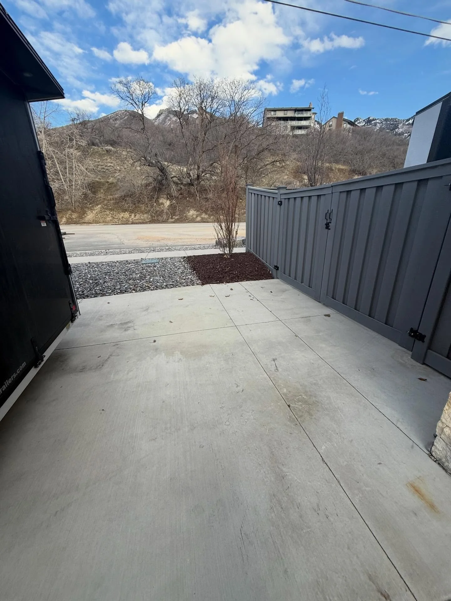 View from a porch or patio showing concrete flooring, a small bush, a gray fence, dirt and gravel area, leafless trees, a road, houses on a hillside, and mountains in the background against a partly cloudy sky.