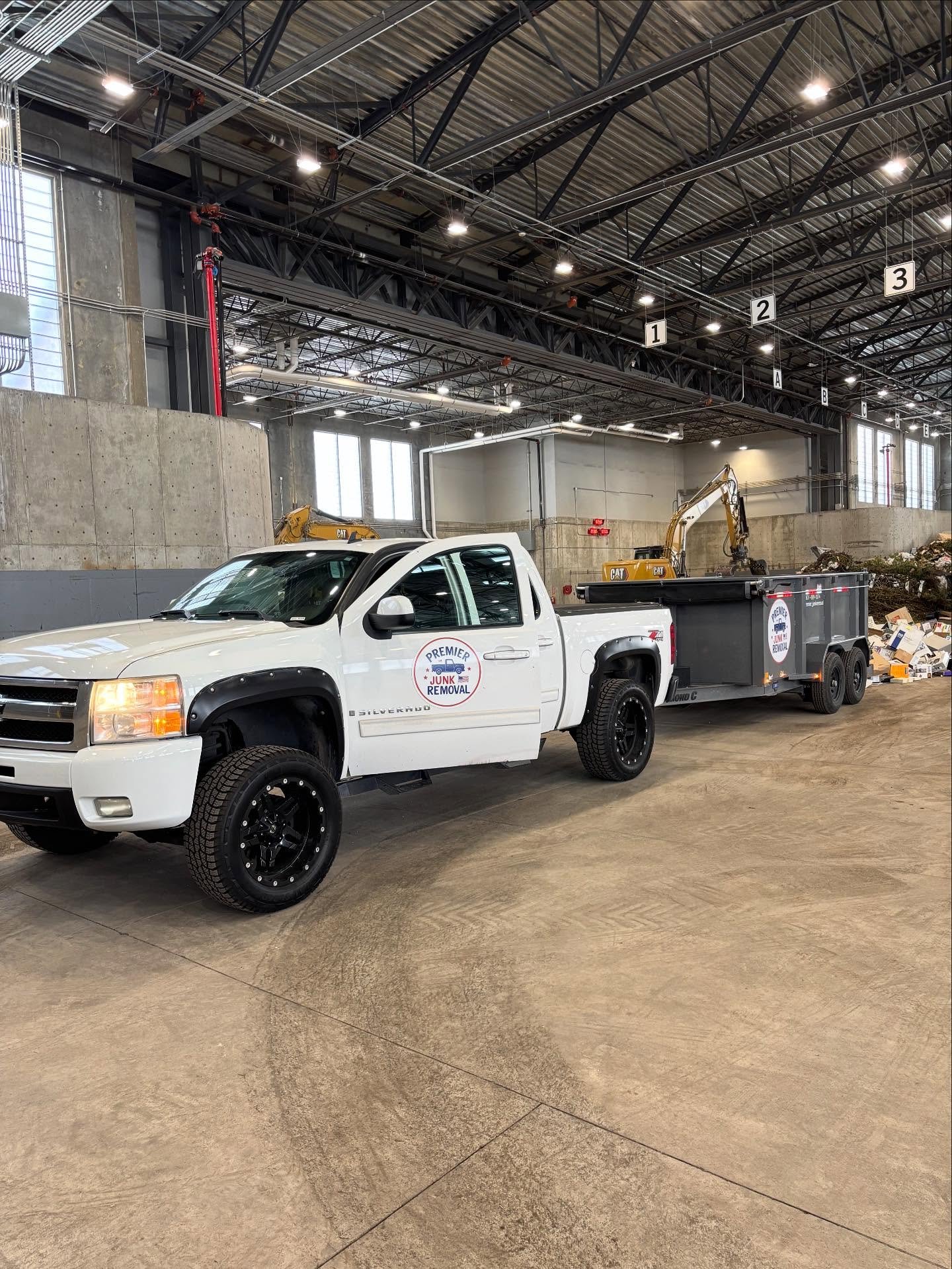 Interior of a large industrial warehouse with a white pickup truck labeled 'Premier Junk Removal' parked inside. The warehouse has high ceilings with visible metal beams, industrial lighting, and a concrete wall. There are small excavators and a large dumpster filled with debris in the background.