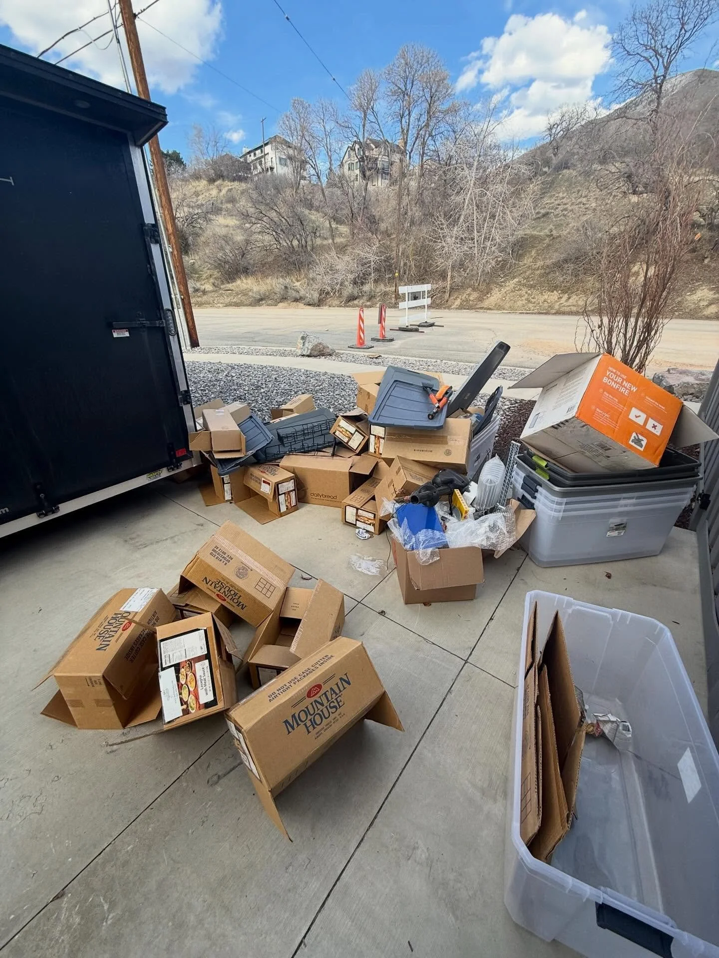 A collection of cardboard boxes, plastic storage containers, and miscellaneous items on a concrete patio outside, with a road, some traffic cones, and a hillside with houses and trees in the background.