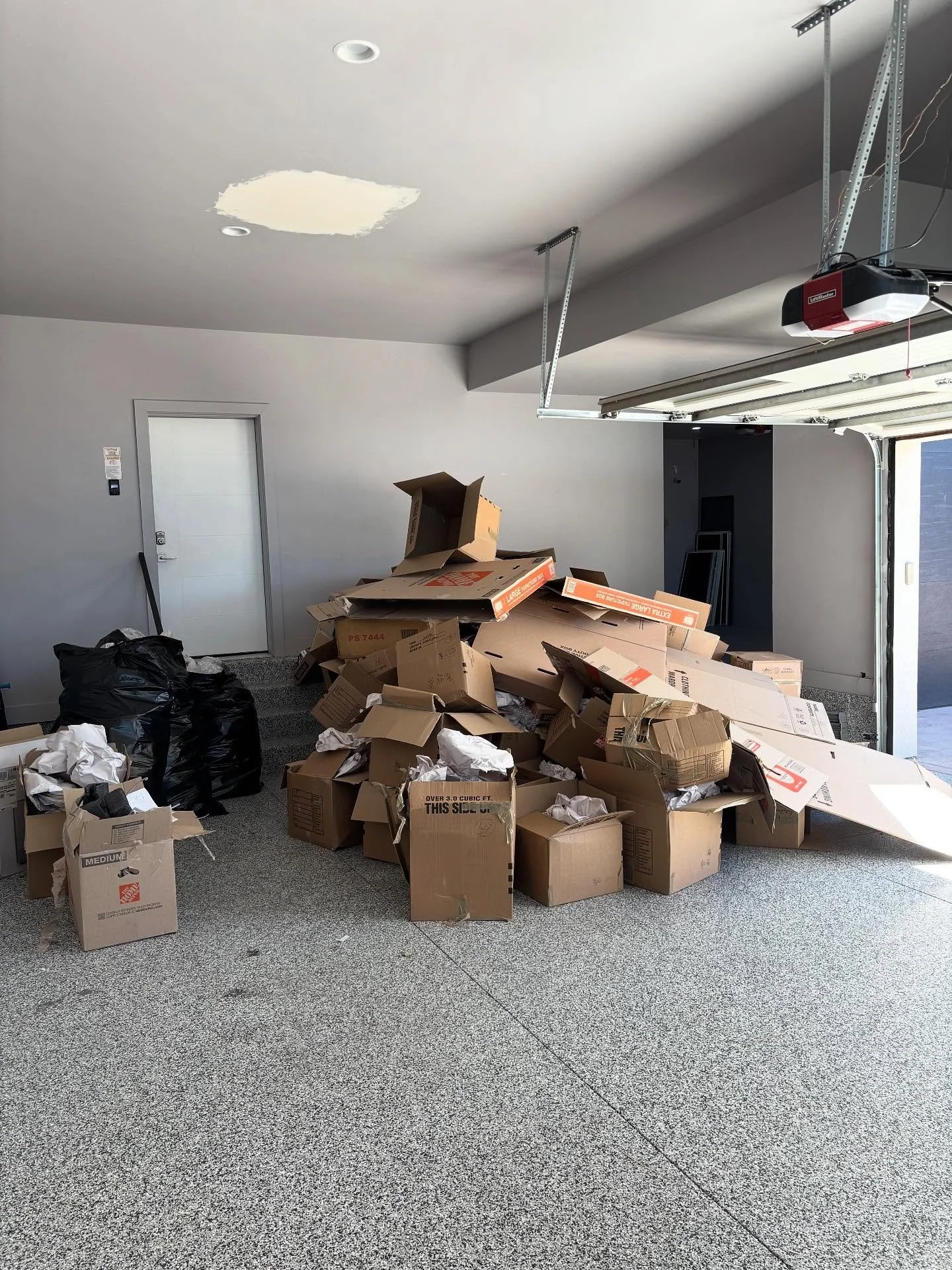 Garage with clutter of empty boxes, some black garbage bags, and crumpled paper on a speckled floor, with a white door, garage door, and ceiling lights.