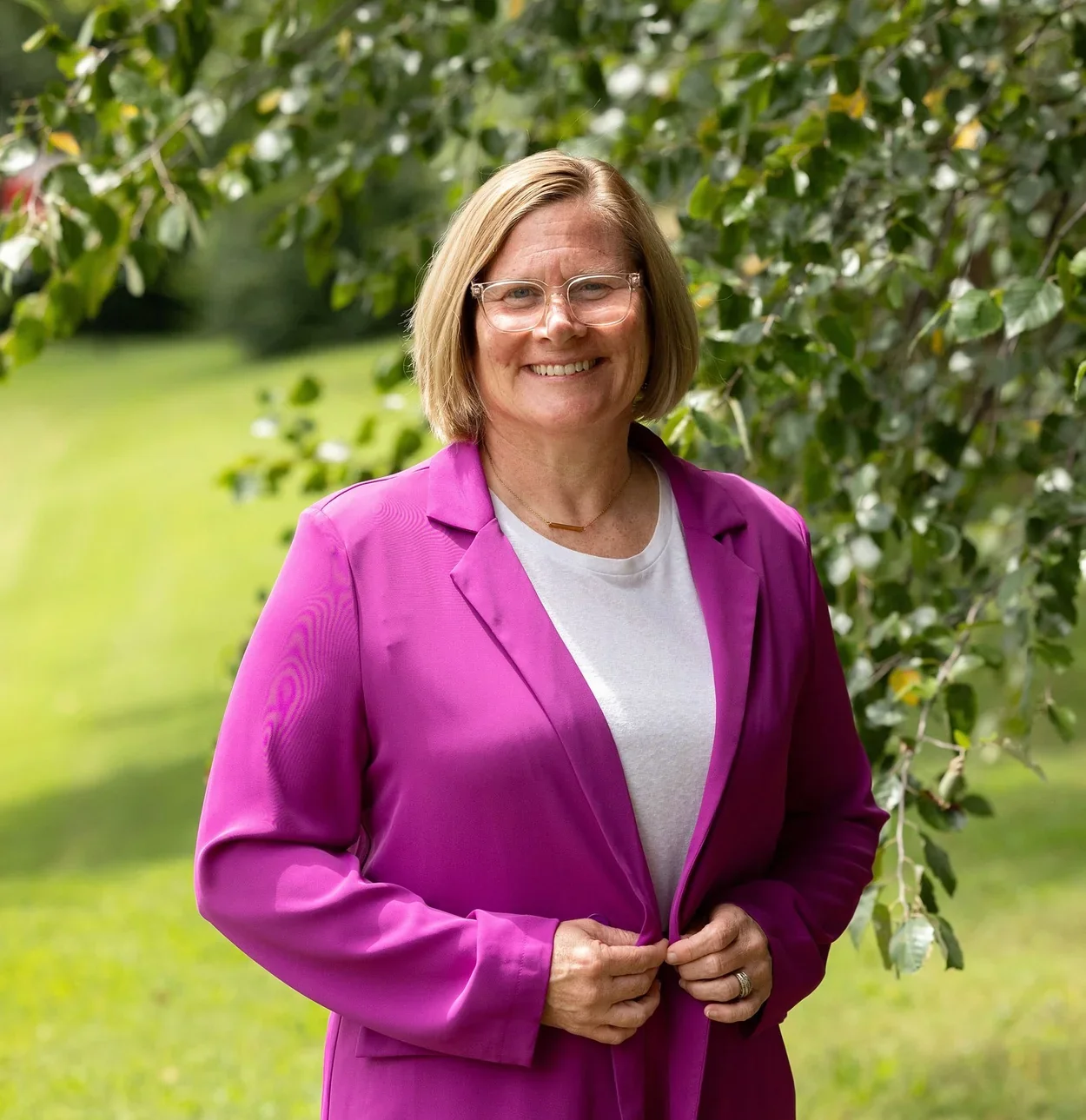 Jen Brooks smiling outdoors in a bright pink blazer, framed by green leaves.